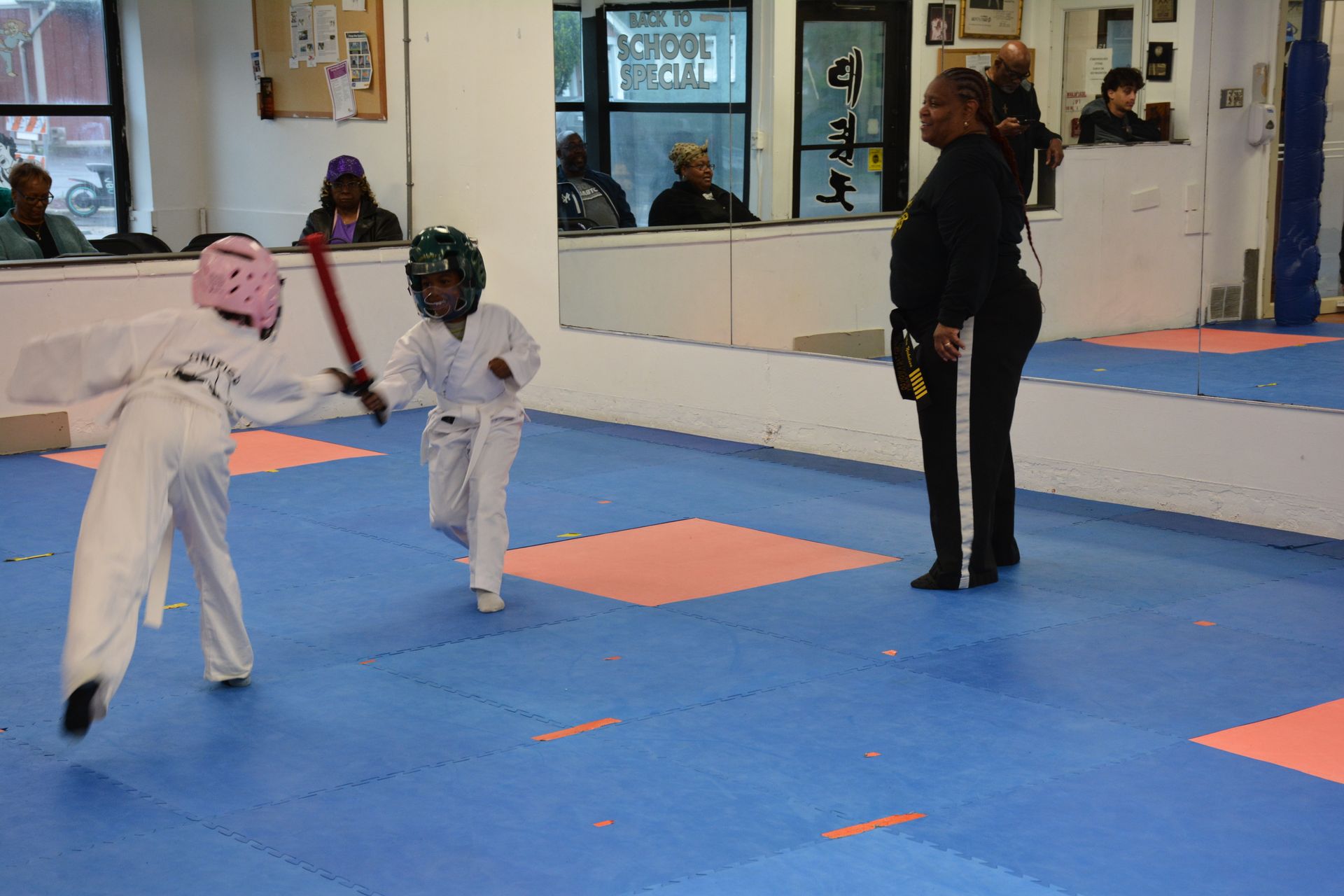 Two children sparring in a martial arts studio on blue mats, with an instructor watching nearby.