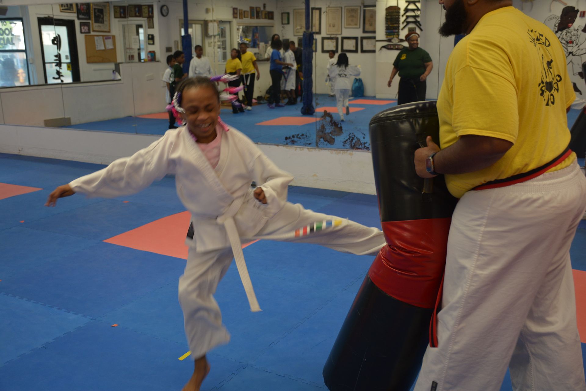 Child in white martial arts uniform kicks a black punching pad in a dojo.