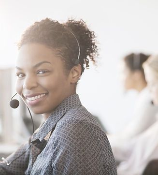 A smiling customer service representative wearing a headset sits in an office with colleagues in the background.