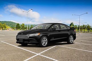 A black Ford Fusion sedan parked in an empty, sunlit paved parking lot with green hills in the distance.