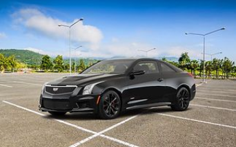 A black Cadillac coupe parked in an empty outdoor parking lot under a bright blue sky with scattered clouds.