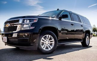 A black Chevrolet Suburban SUV parked on an asphalt lot under a bright blue sky.