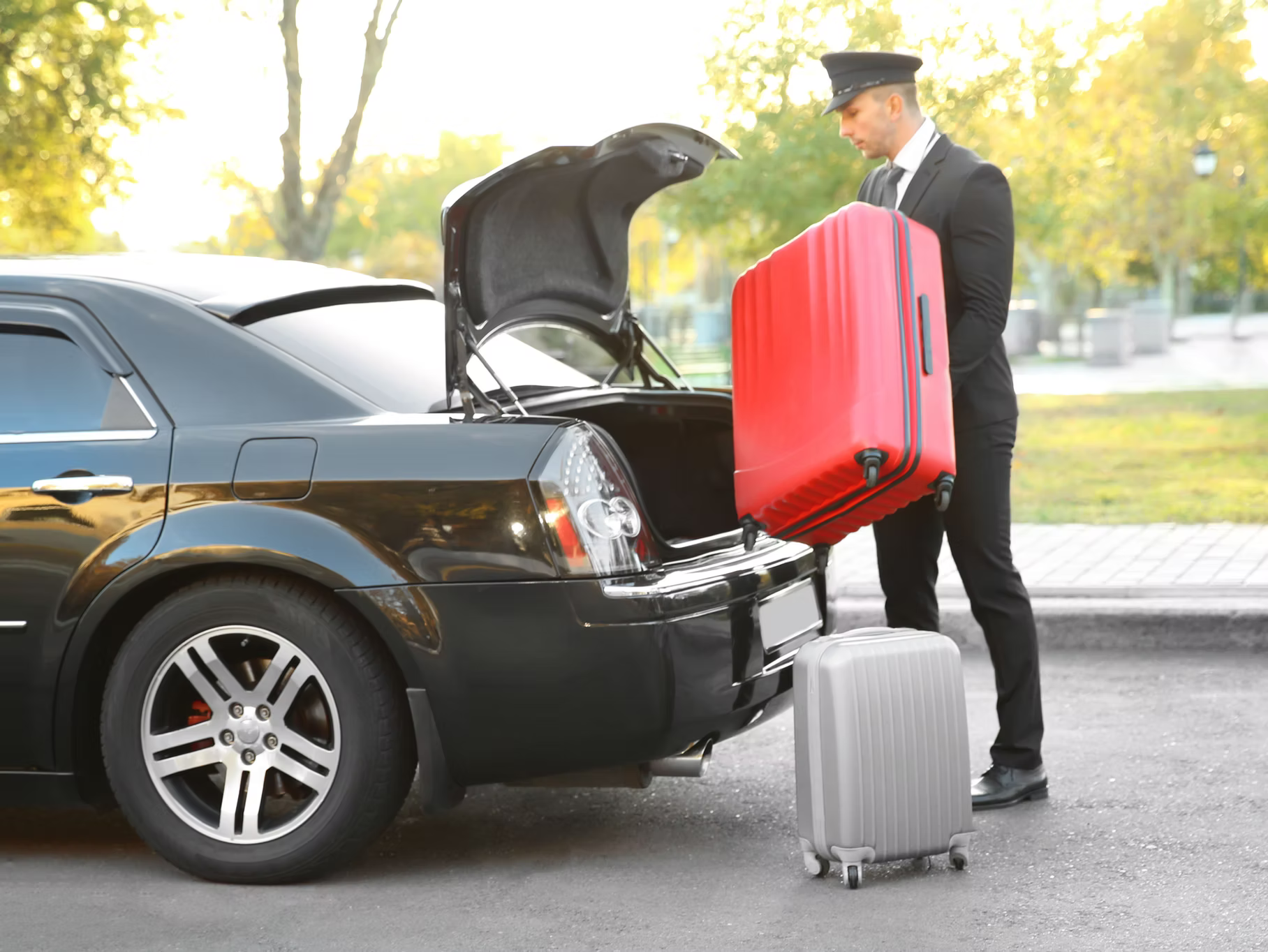 A chauffeur in a suit loads a large red suitcase into the open trunk of a black car, with a silver suitcase on the ground.