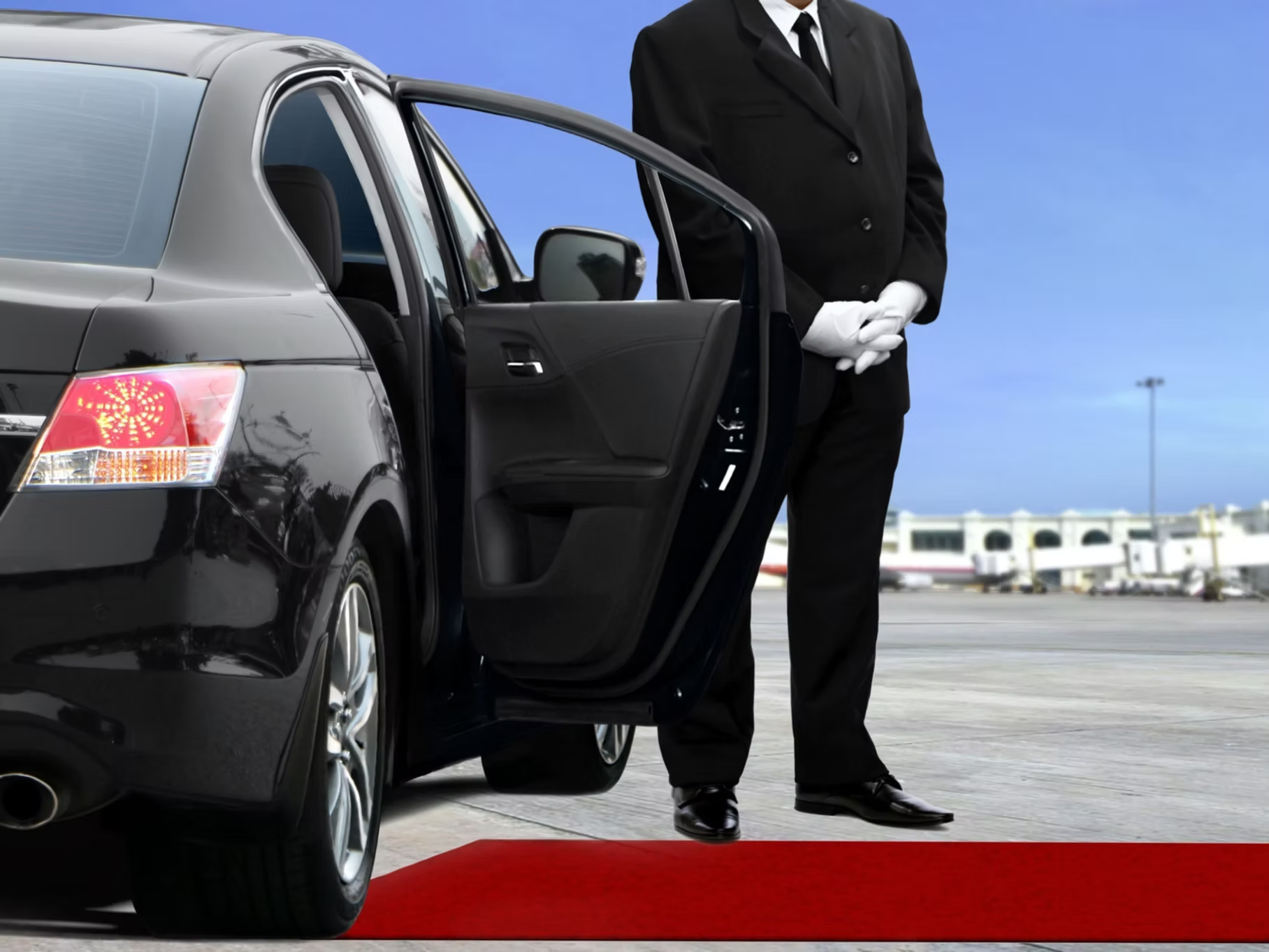 A chauffeur in a suit and white gloves stands by an open car door next to a red carpet at an airport.