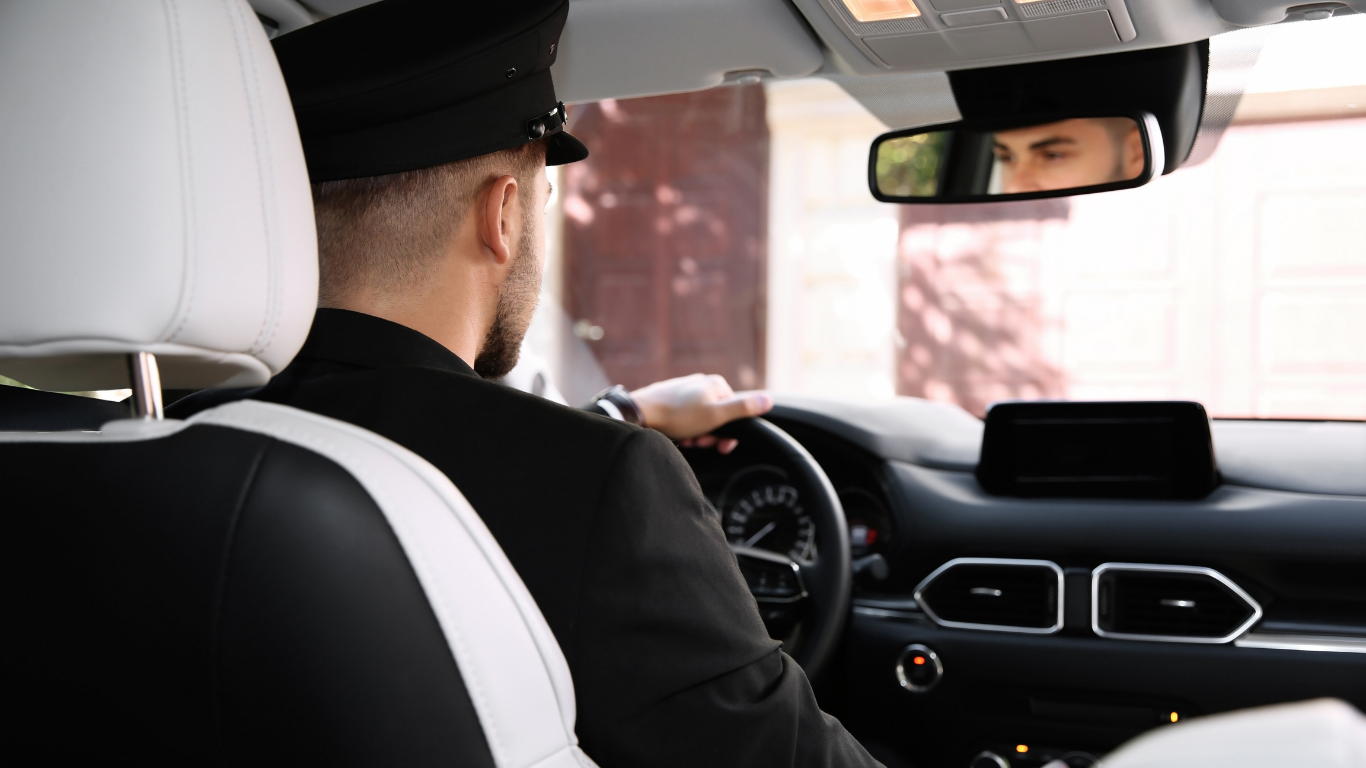 A person in a black uniform drives a car, seen from the back seat. Their face is visible in the rearview mirror.