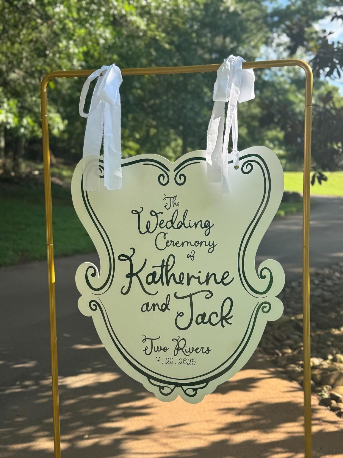 Decorative wedding sign hanging outdoors for Katherine and Jack, with white ribbons and green lettering.