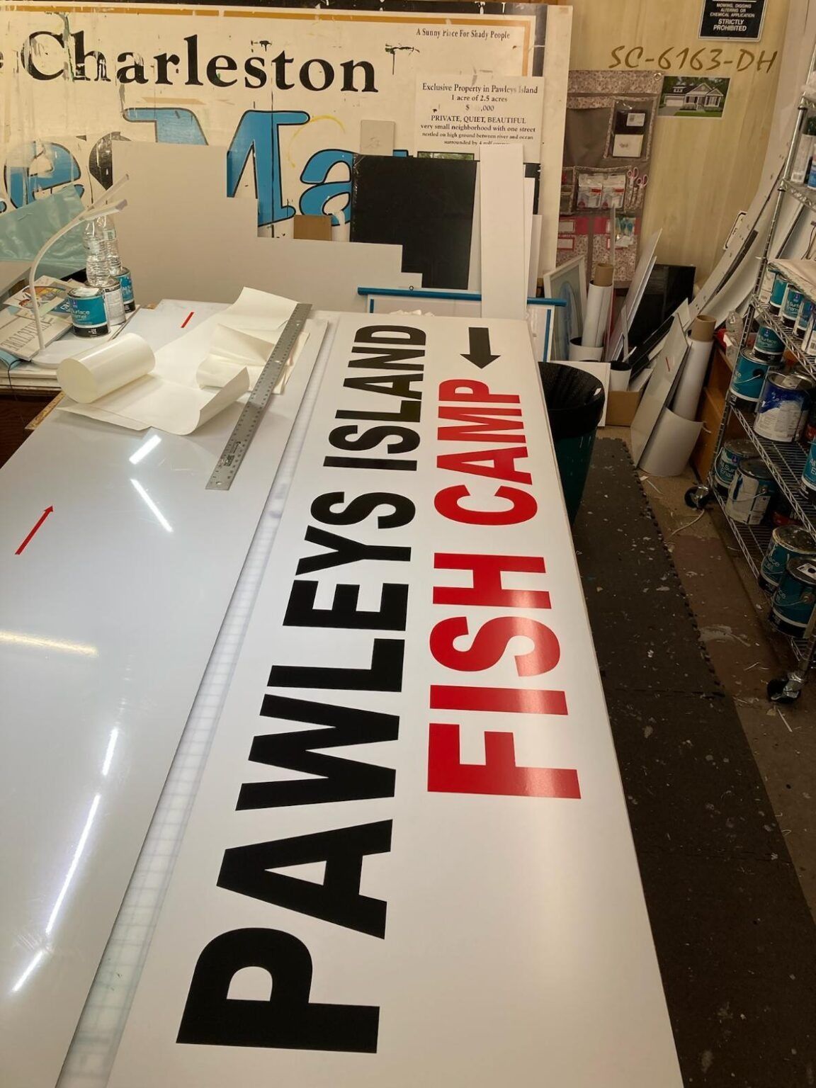 Large sign reading “PAWLEYS ISLAND FISH CAMP” in a workshop, with Charleston sign elements nearby.