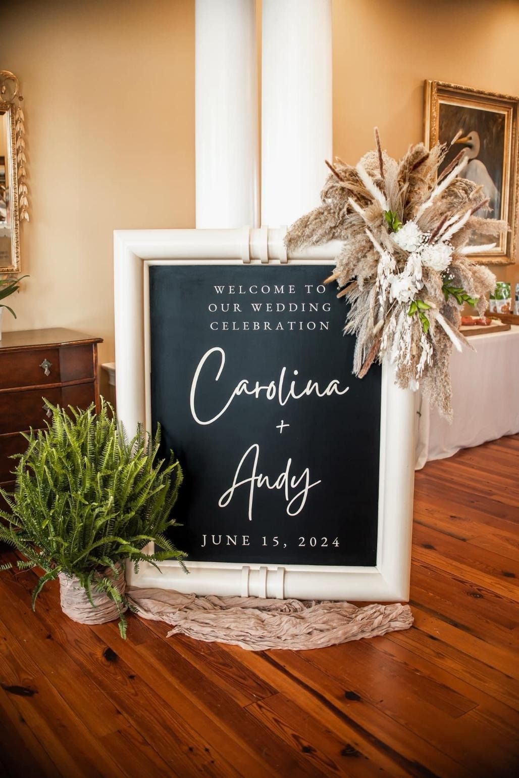 Wedding welcome sign on chalkboard easel with white frame, greenery, and dried floral arrangement in a warm indoor venue