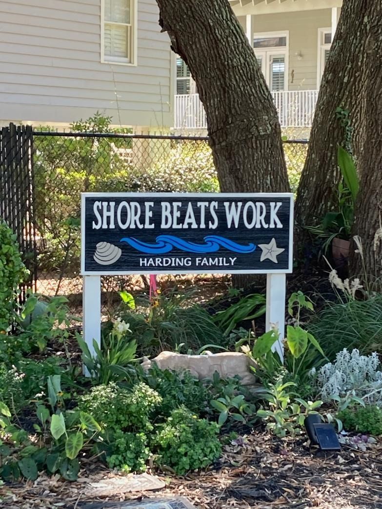 Shore Beats Work sign in a garden bed outside a house, with flowers and greenery.