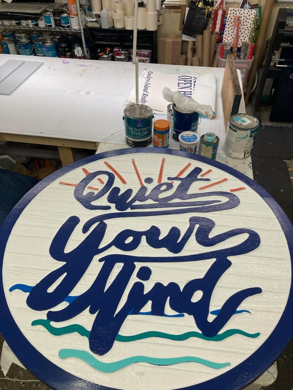 Round sign reading “Quiet your mind” in blue script with teal waves and orange rays, in an art studio