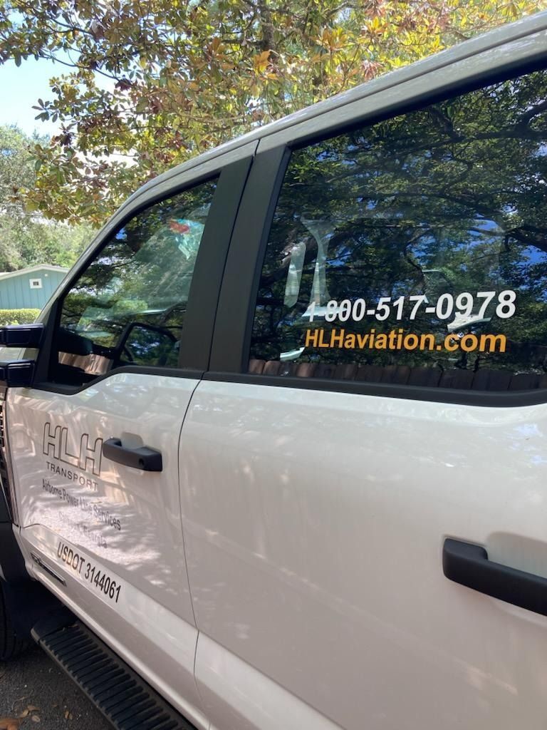 White van with HL Aviation.com and a phone number on the side, parked outdoors under trees.