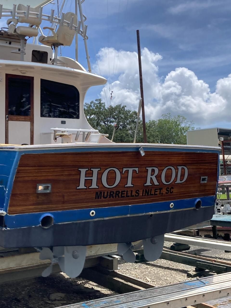 Close-up of a wooden boat stern on a trailer, labeled “HOT ROD,” under a bright cloudy sky.