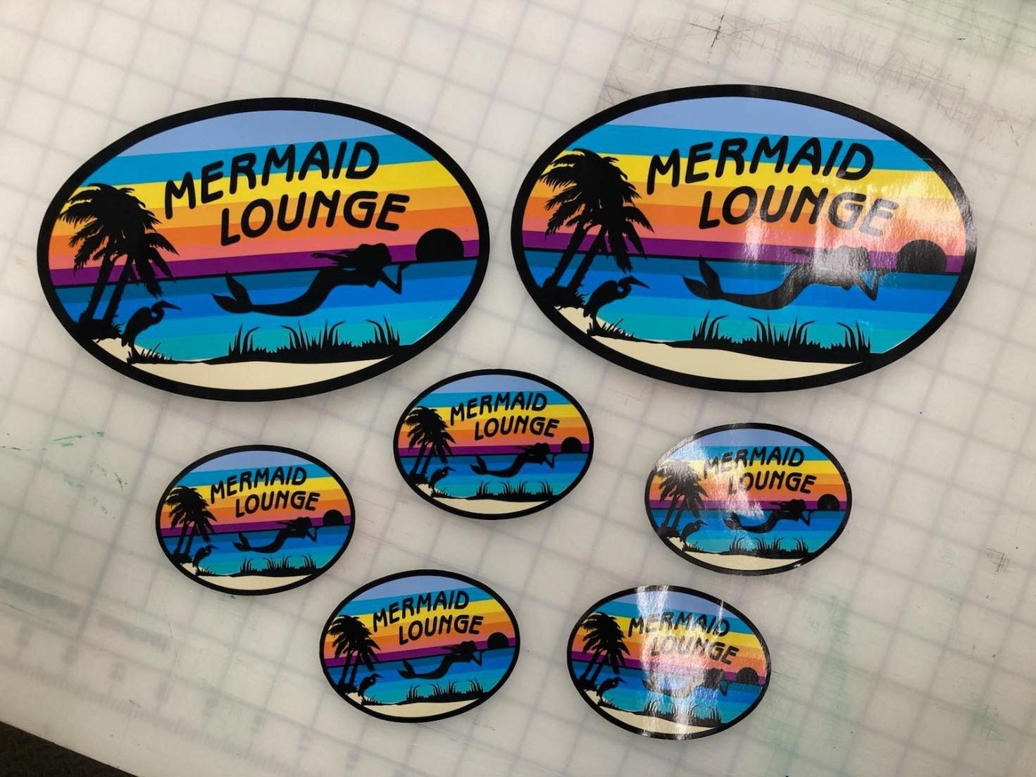Six oval Mermaid Lounge stickers with sunset beach and palm tree designs on graph paper