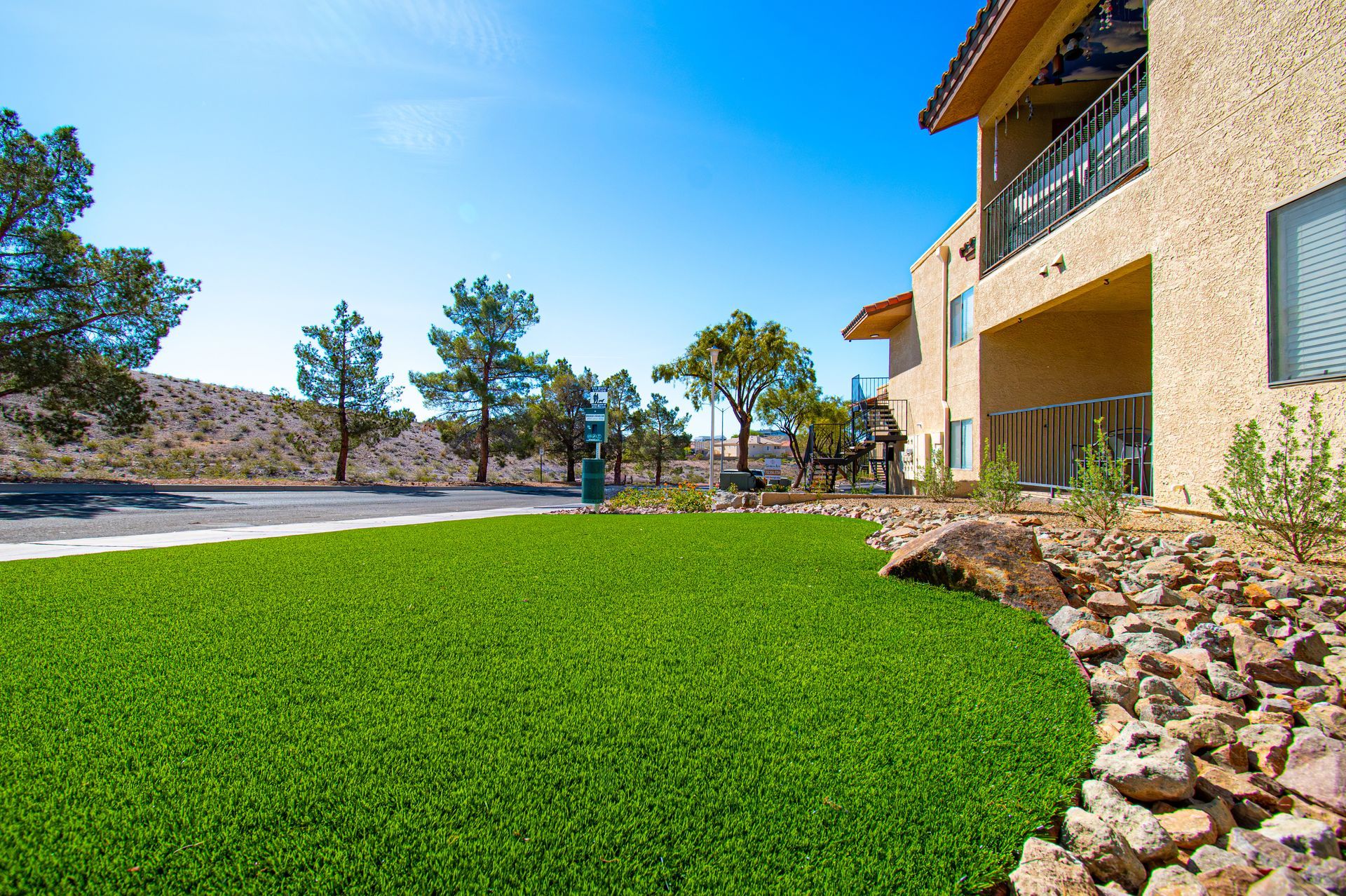 Landscaped yard with green lawn, rocks, trees, and a stucco house under a bright blue sky