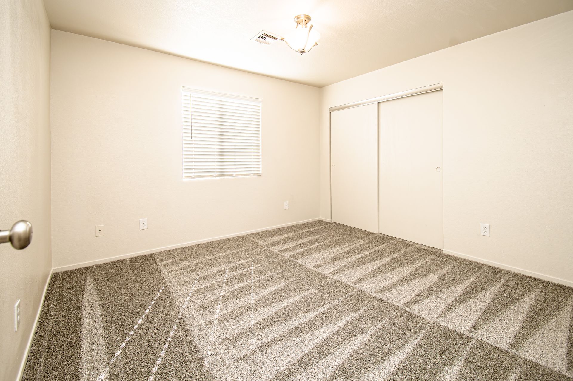 Empty beige bedroom with carpeted floor, ceiling fan, window blinds, and closet doors
