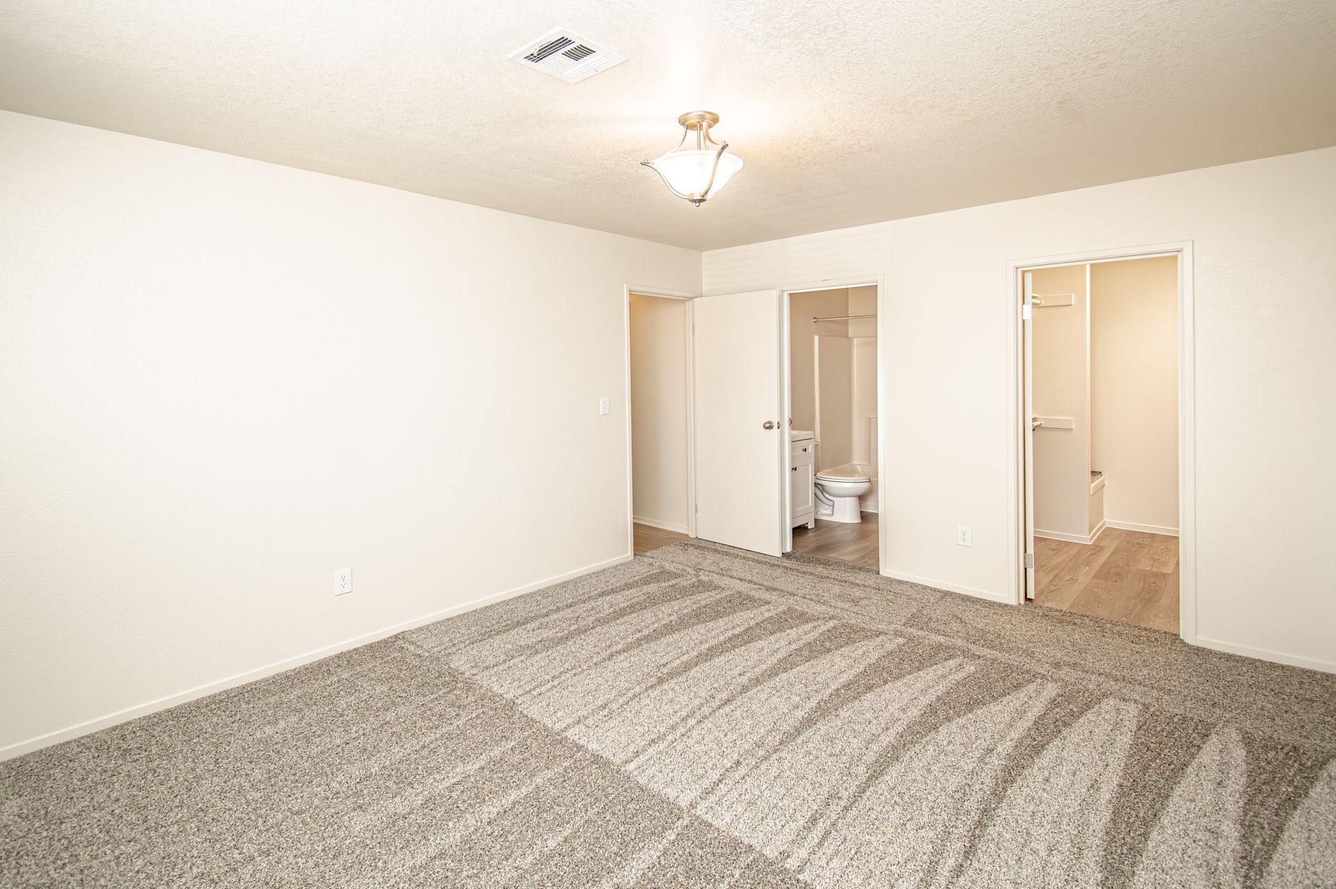 Empty carpeted bedroom with white walls, ceiling light, and open doors to a bathroom and closet.