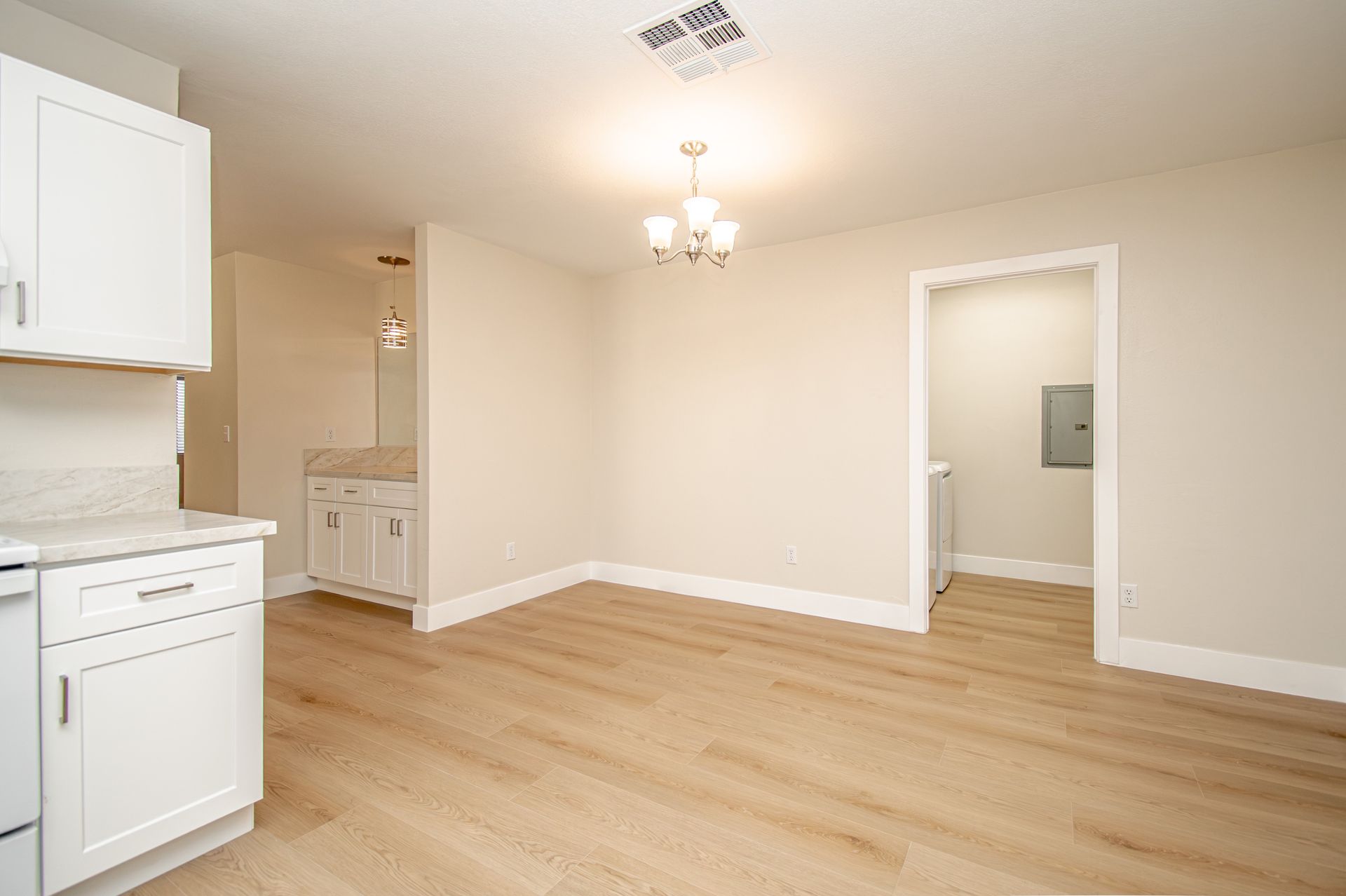 Empty beige kitchen with white cabinets, light wood flooring, and a doorway to another room