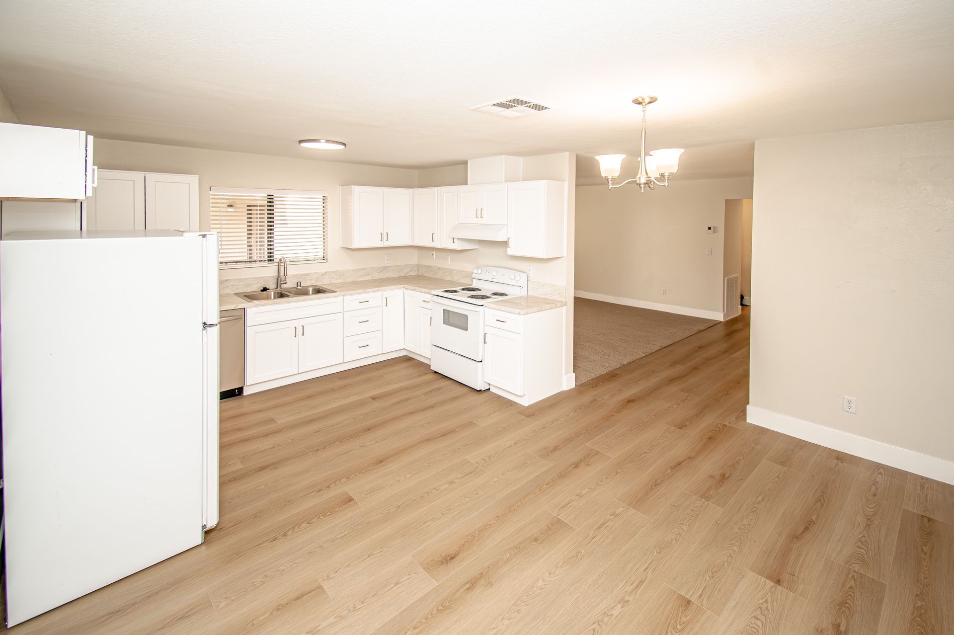 Bright empty kitchen with white cabinets, refrigerator, and light wood floors opening into a living area