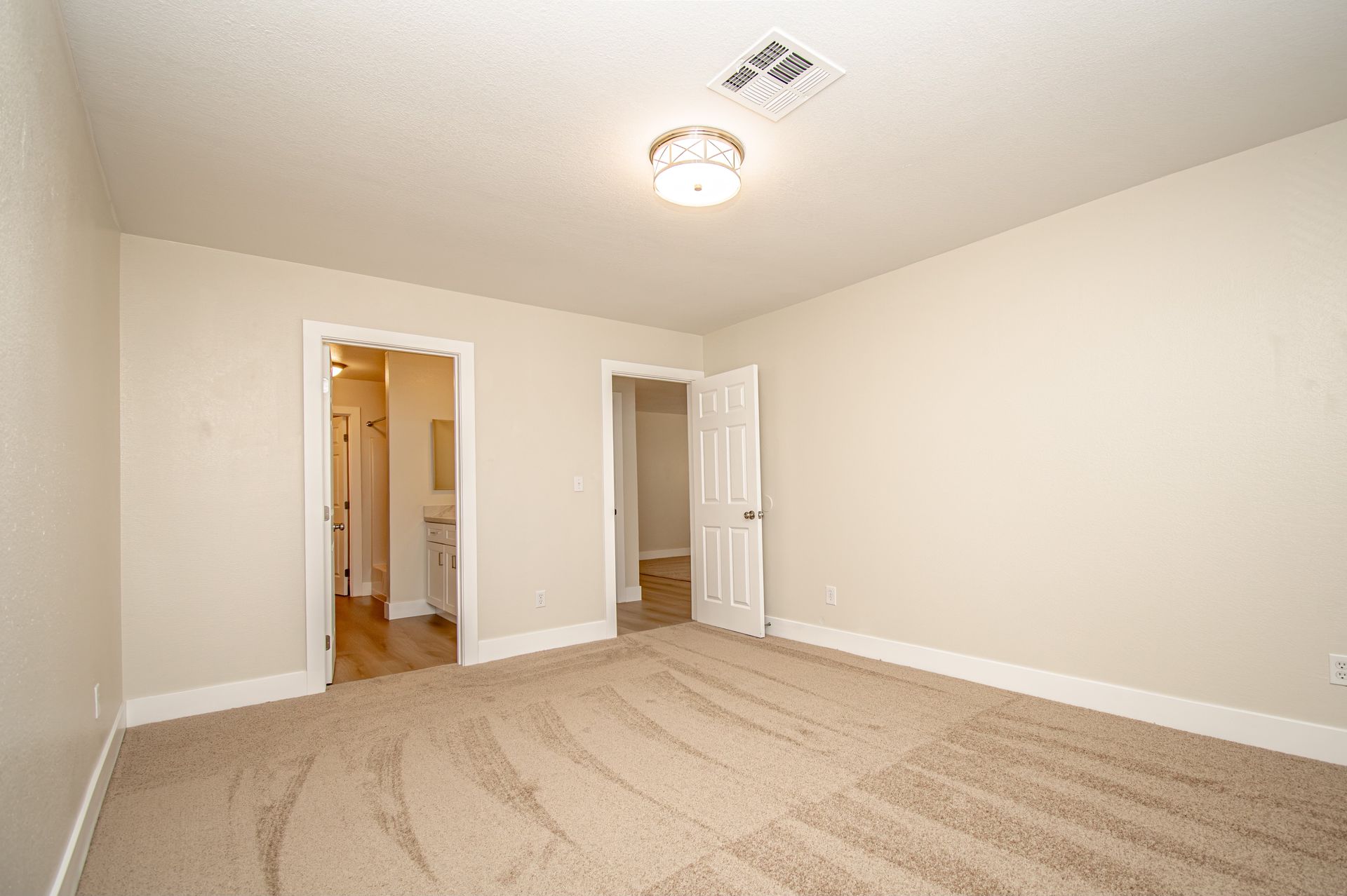 Empty beige carpeted bedroom with white walls, ceiling light, and two open doorways.