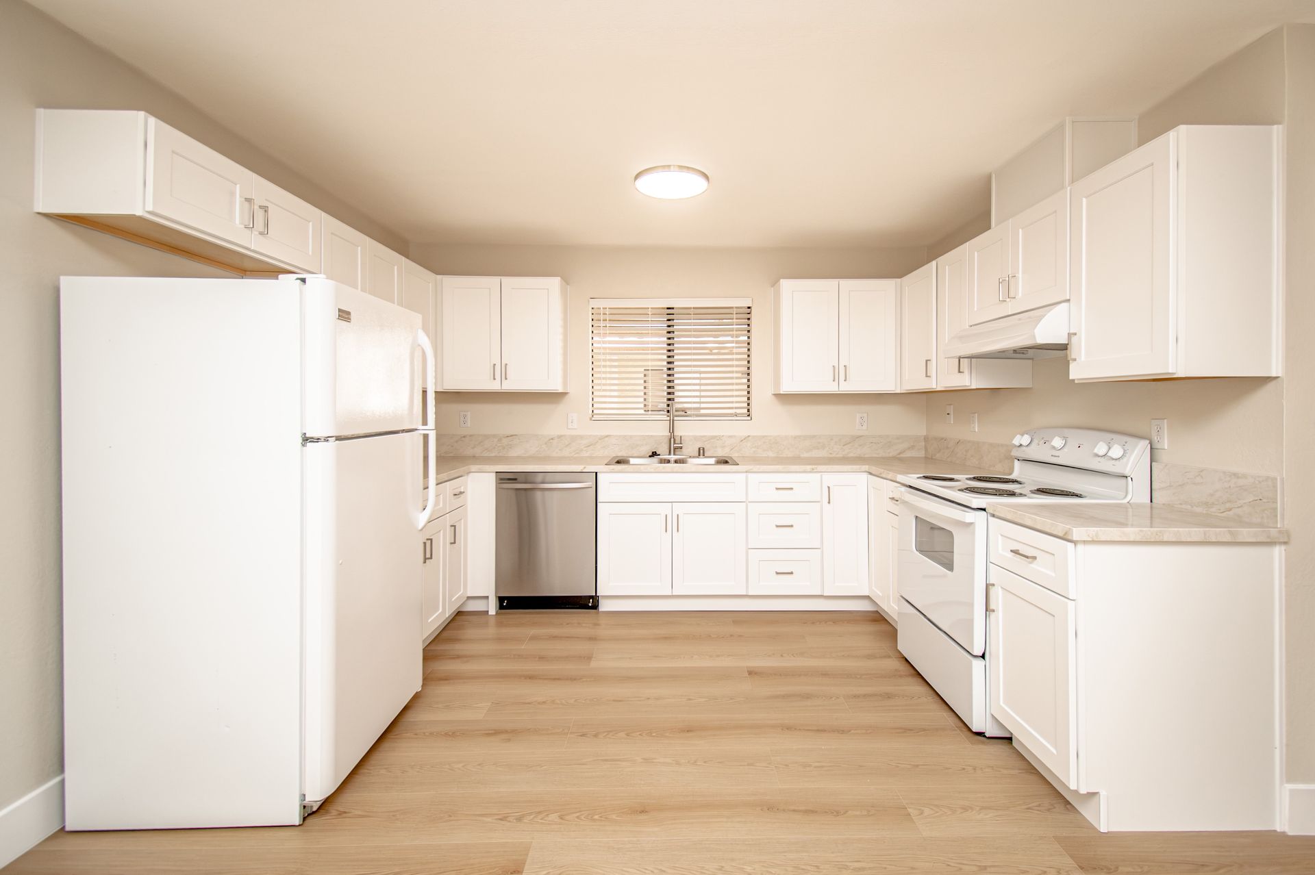 Bright white kitchen with wood floors, white cabinets, refrigerator, stove, and sink under a window.