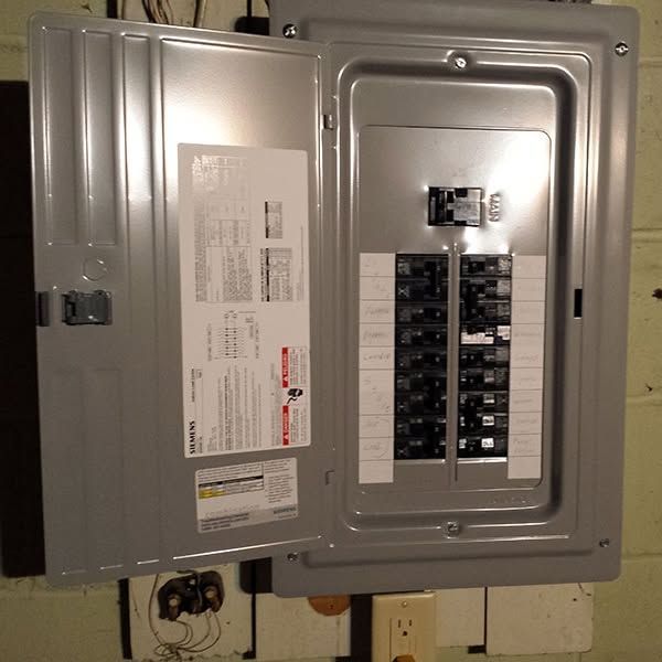 Electrician kneeling beside an open electrical panel, testing wires with a multimeter in a workshop.