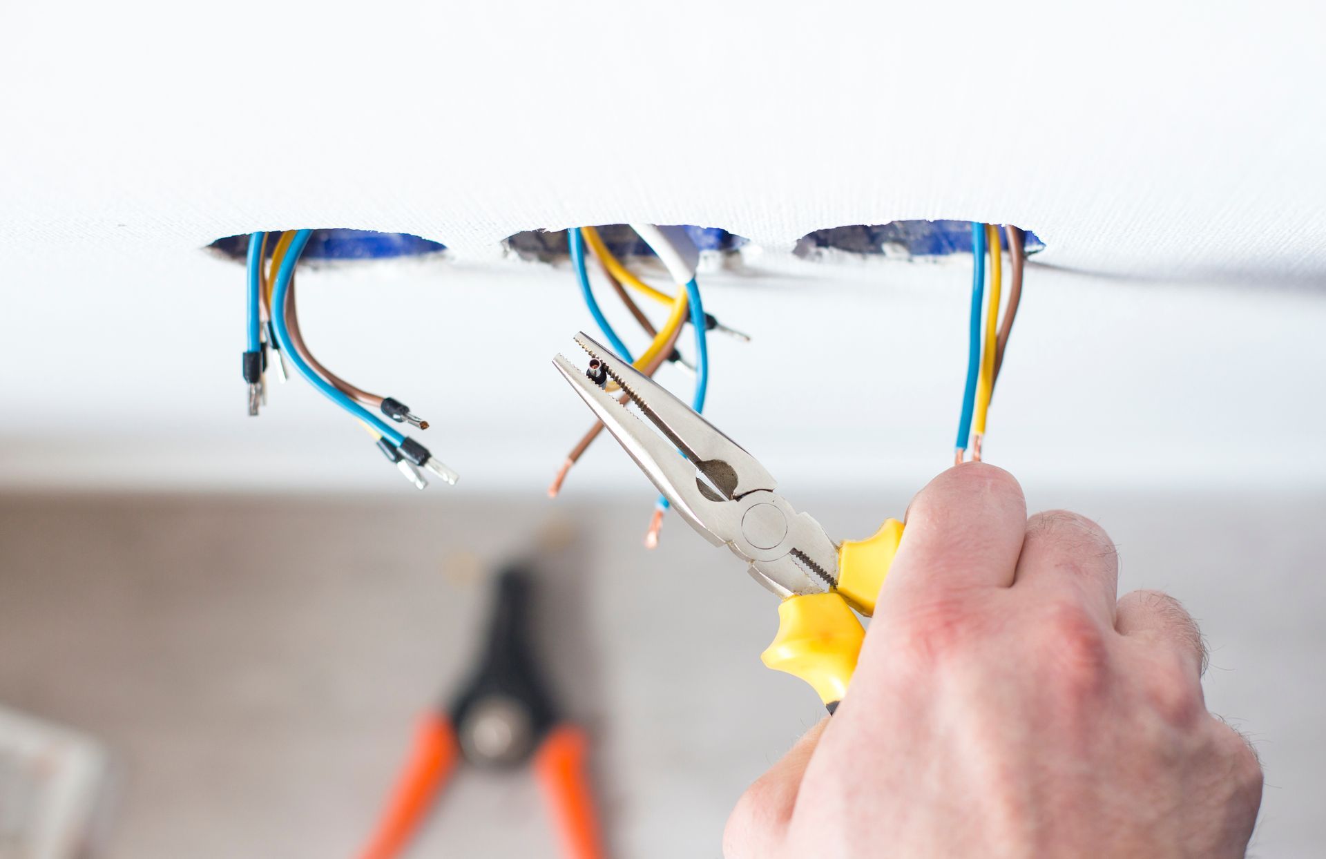 Hand cutting electrical wires in a wall opening with pliers, exposing blue, black, and yellow cables.