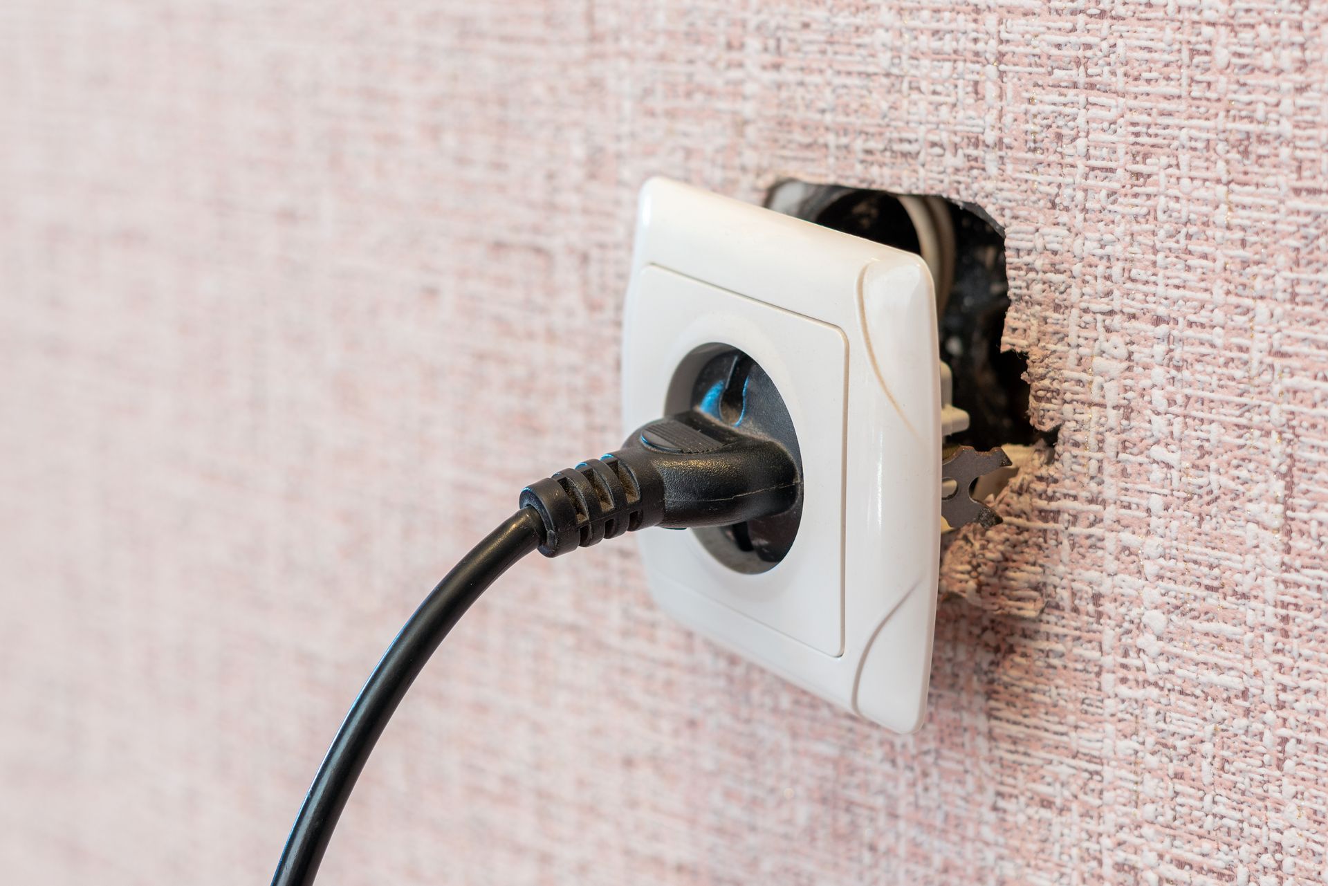 Black power plug in damaged wall outlet on beige textured wall.