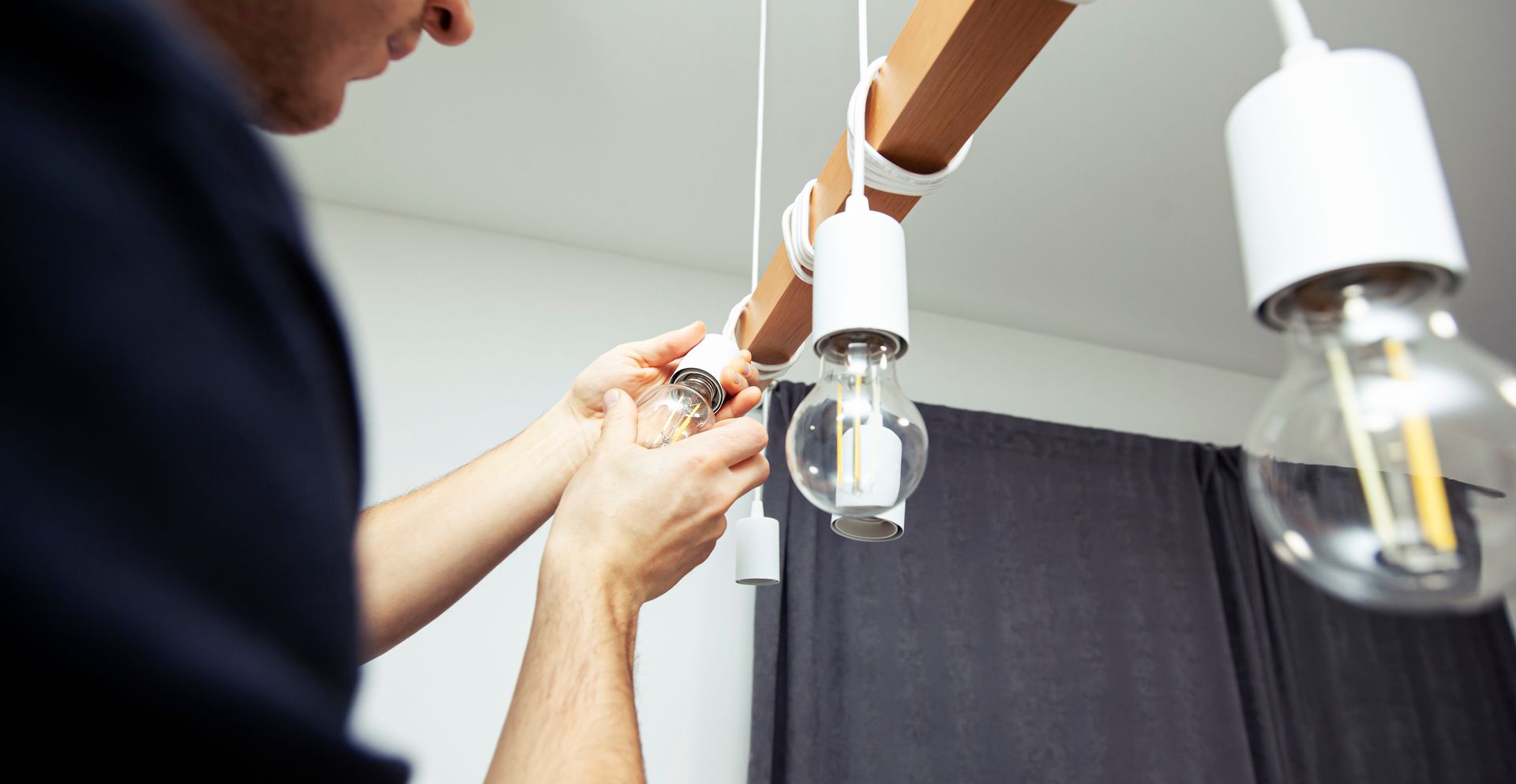 Person installing a hanging light bulb fixture indoors beside dark curtains.