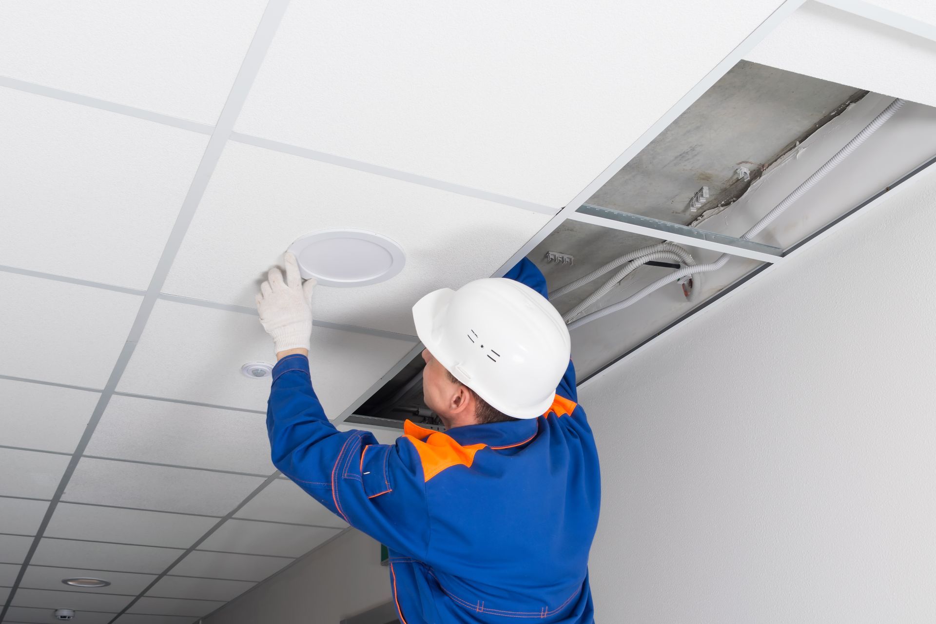 Worker in blue uniform installing a ceiling light panel in an office ceiling.