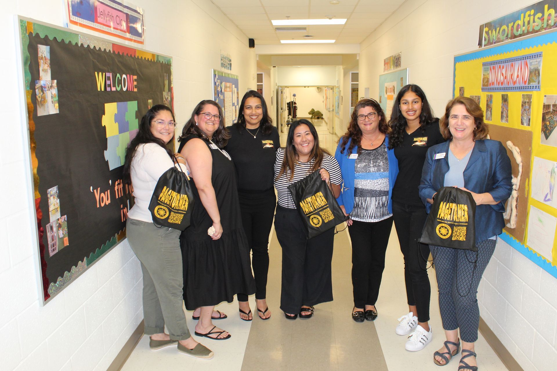 Group of teachers in a school hallway, holding bags. They are smiling at the camera.