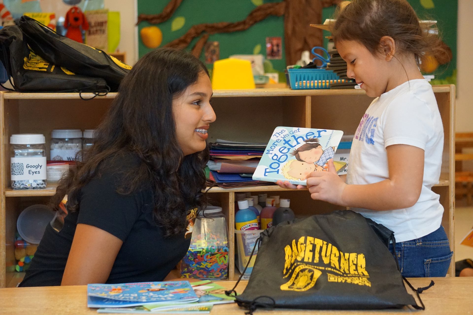 Woman and child reading a book together at a desk in a classroom. The woman is smiling.