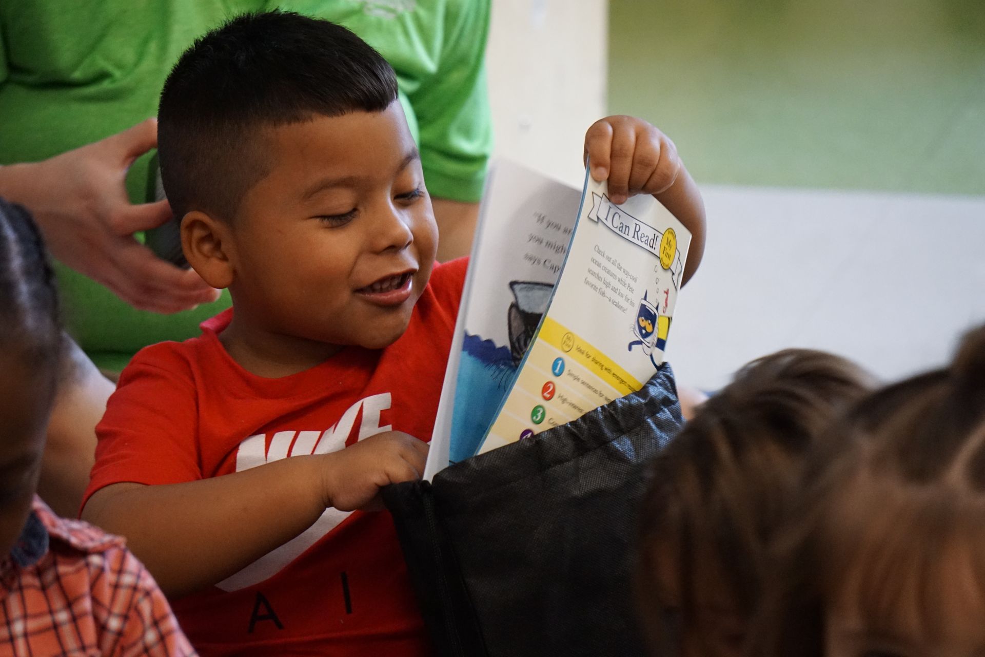 Young boy in red shirt smiles, holding a book, appears to be reading.