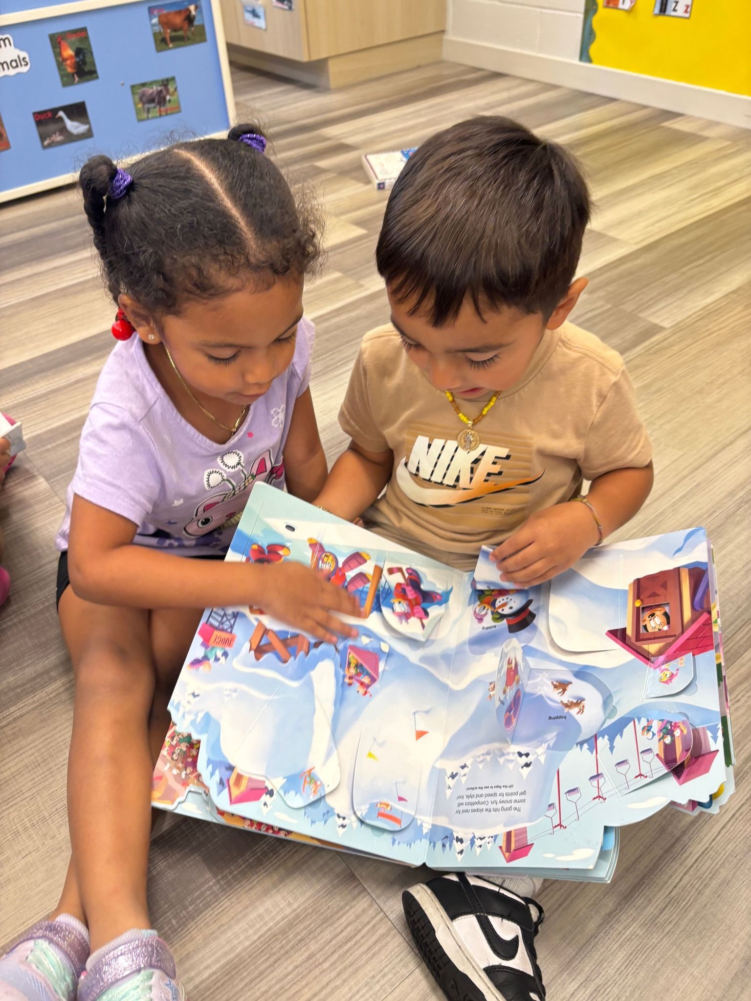 Two young children seated, looking at a colorful storybook with pop-ups, indoors.