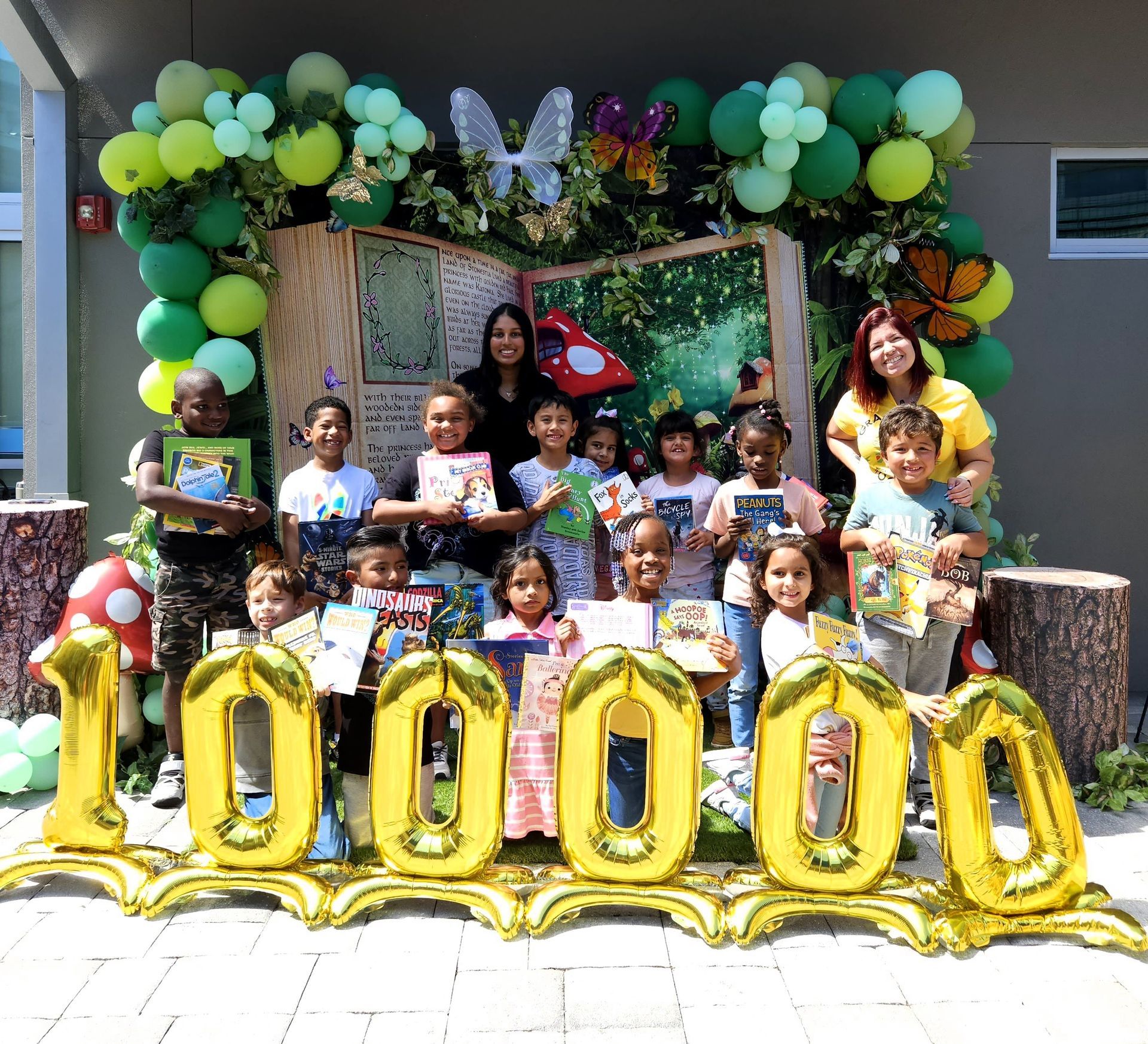 Children and adults celebrate a milestone with books. Gold balloons spell 1000. Green and white balloon arch.