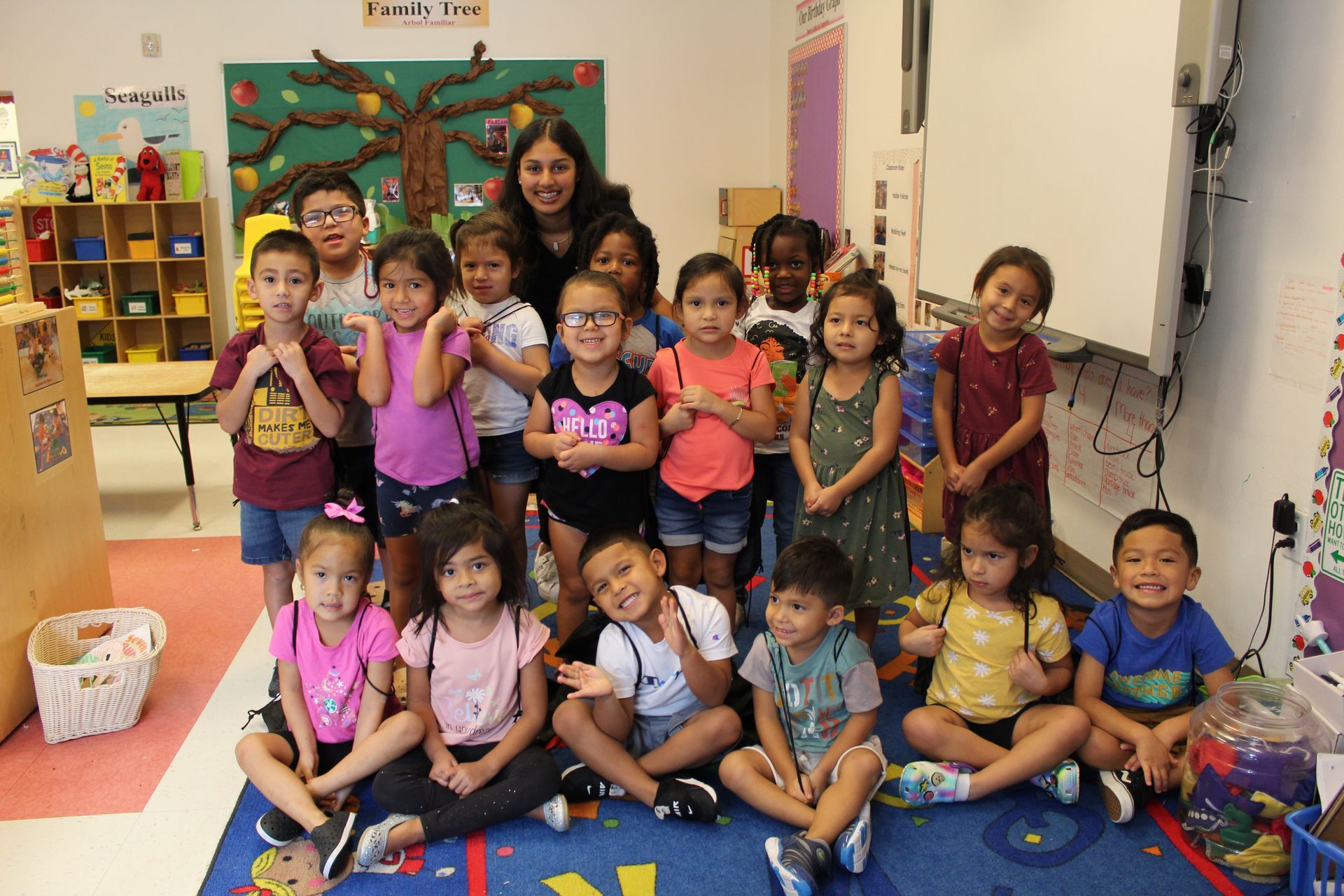 A group of children and a teacher smiling in a colorful classroom.