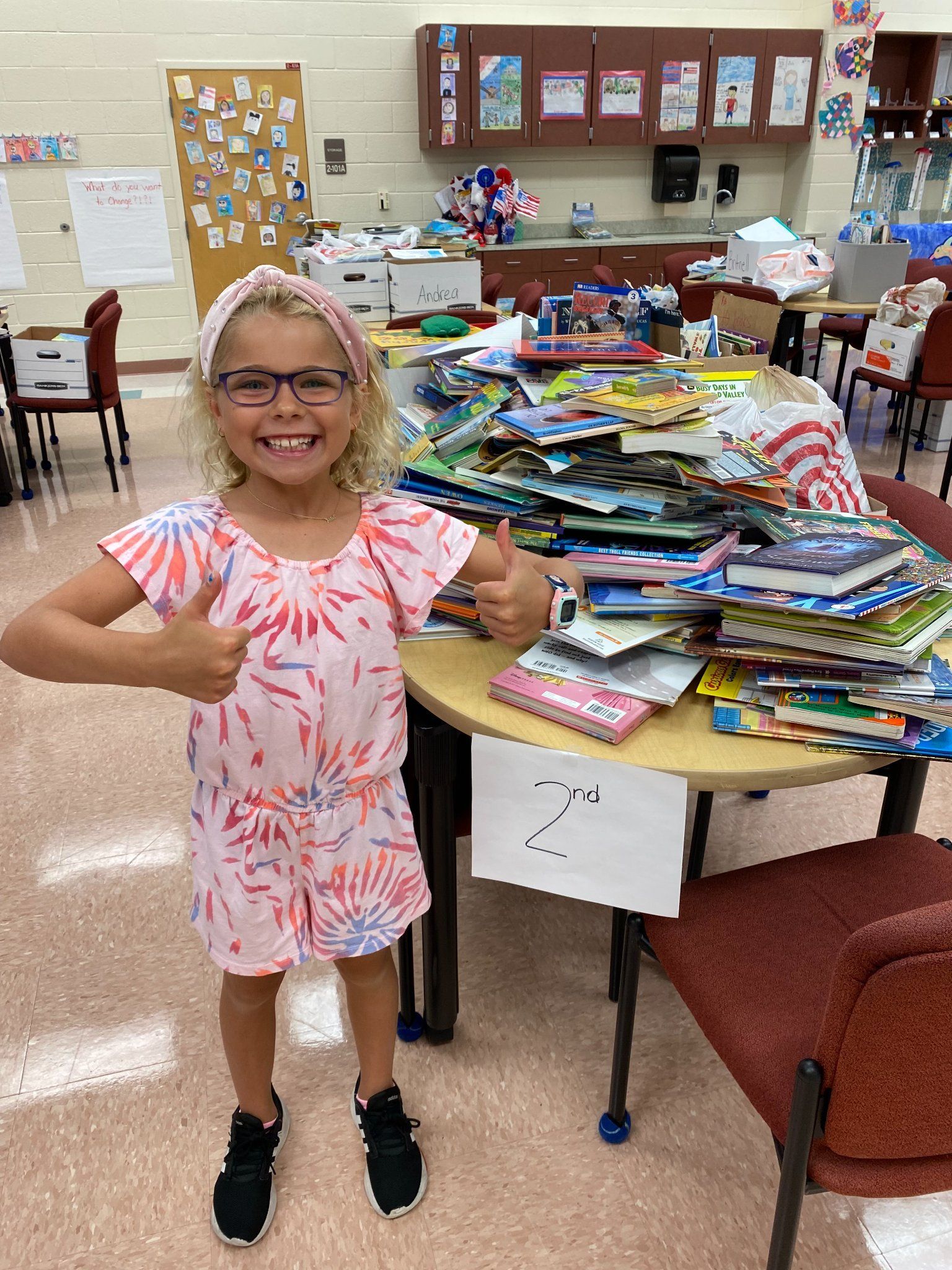Girl gives thumbs up next to a table piled high with books in a classroom.