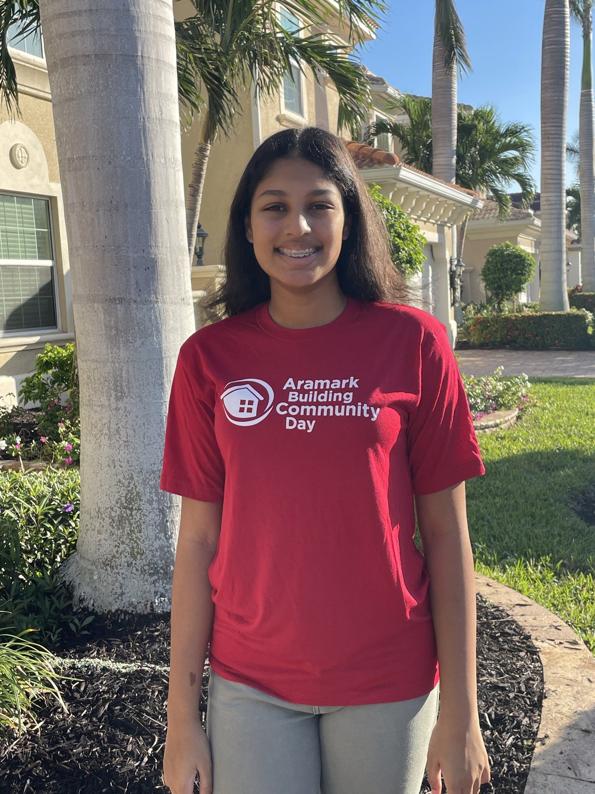 Young woman wearing a red t-shirt standing outside, smiling. Background includes a house and palm trees.