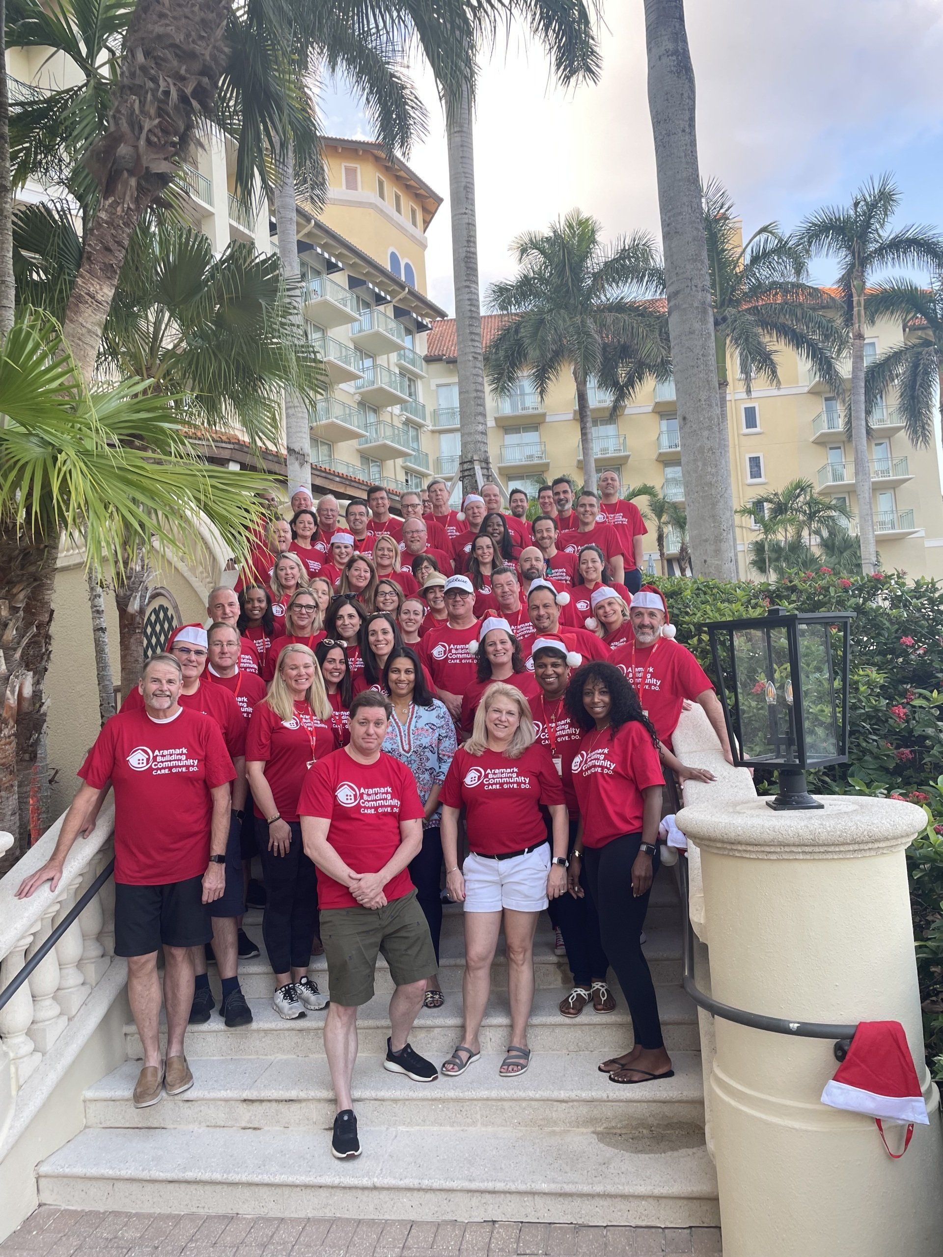 Large group in red shirts on steps; sunny outdoor setting near building; some wearing Santa hats.