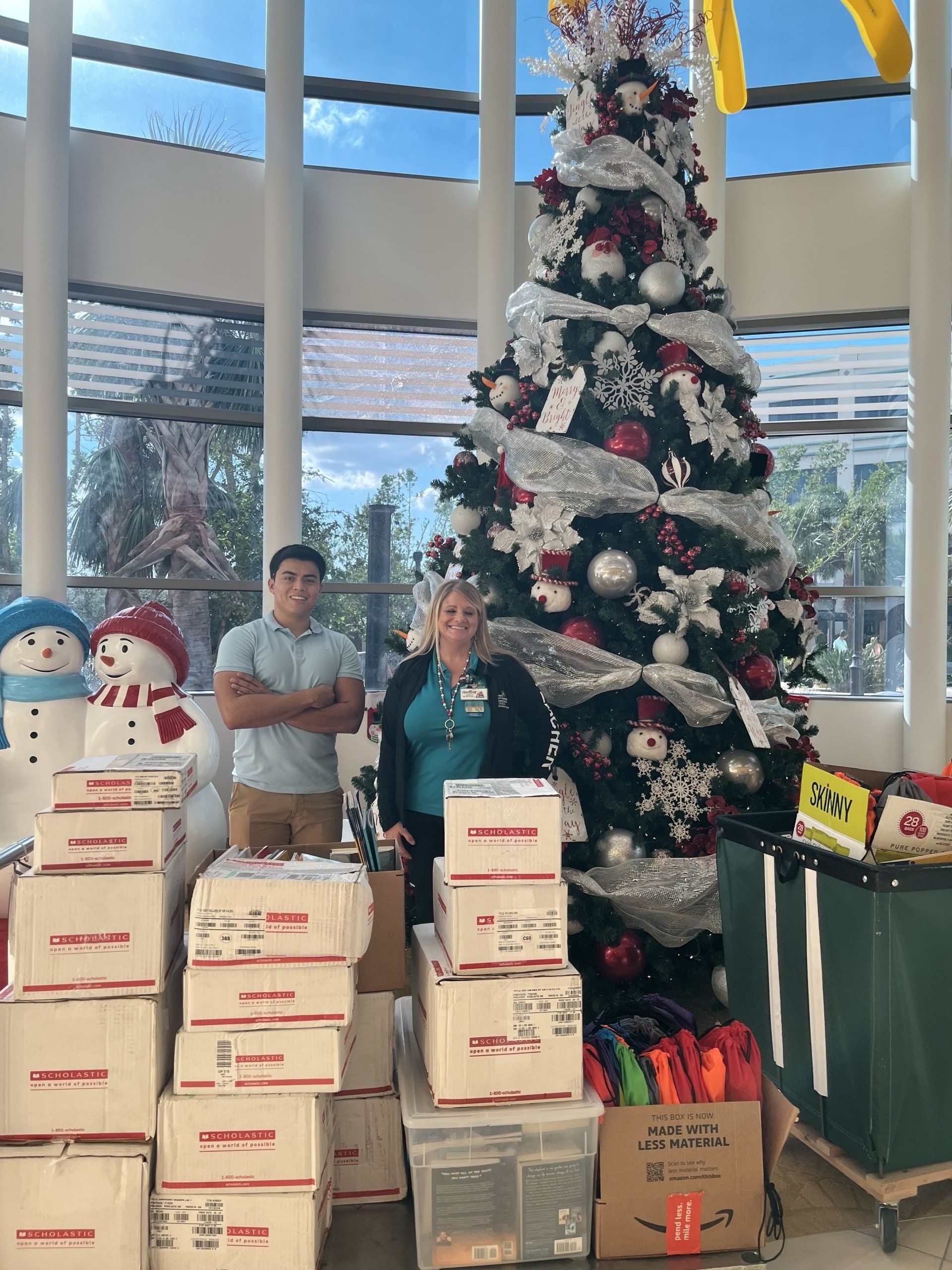 Two people pose with gifts beside a decorated Christmas tree in a lobby. Boxes and gifts are stacked.