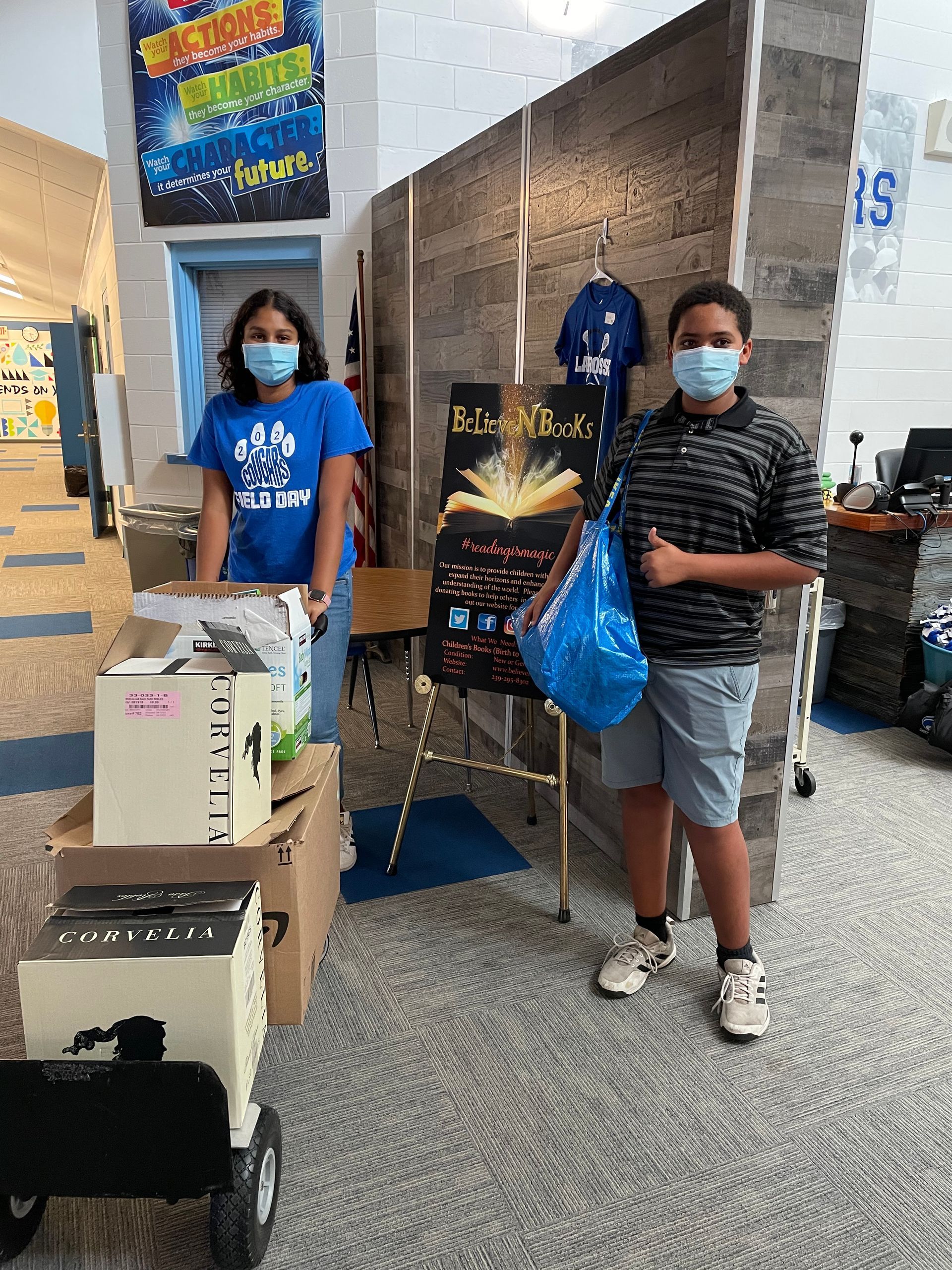Two students wearing masks, selling items. One holds a bag, the other stands behind a cart with boxes and a sign.