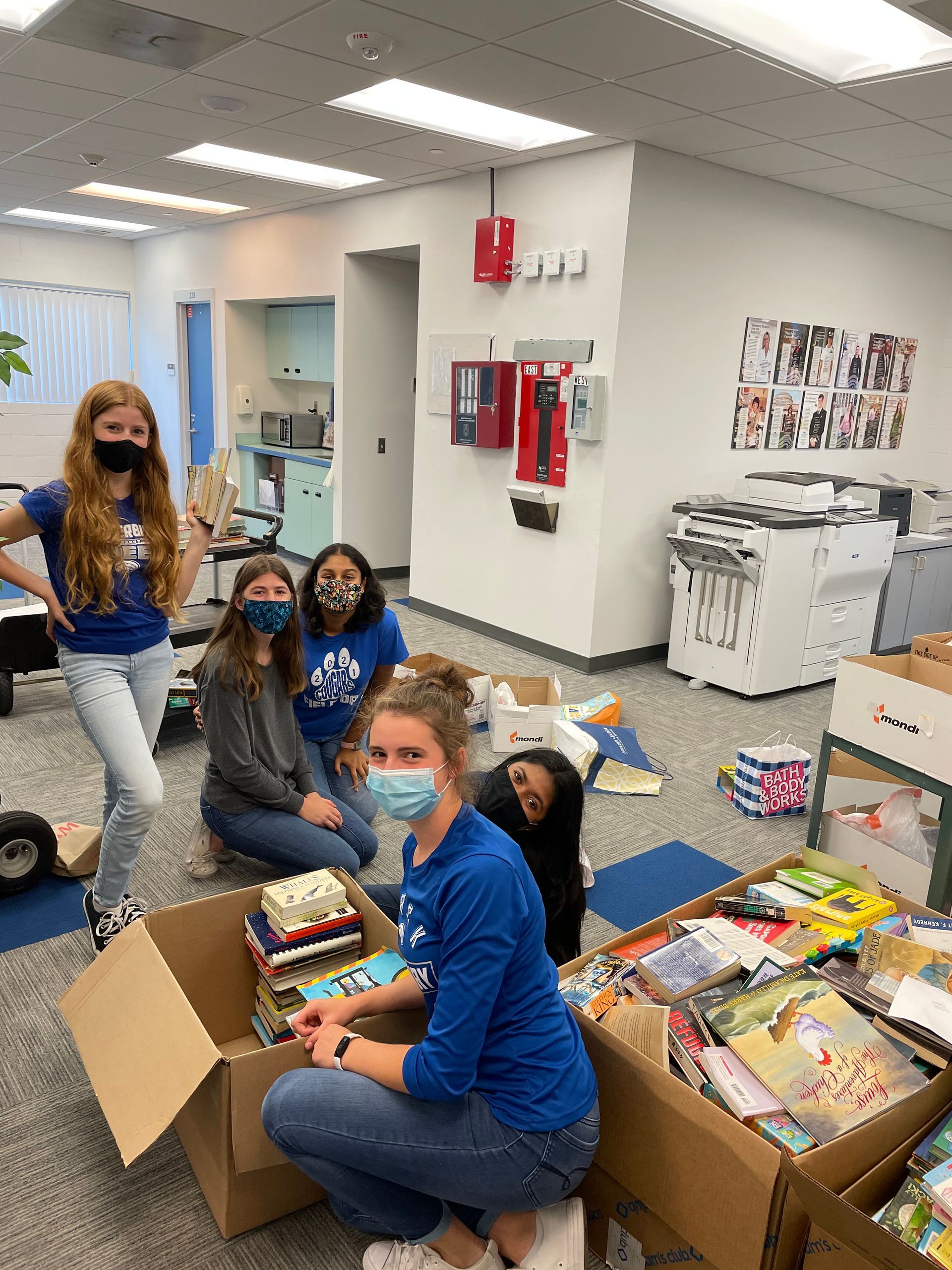 Five people in blue shirts sorting books into boxes in a room with printers.