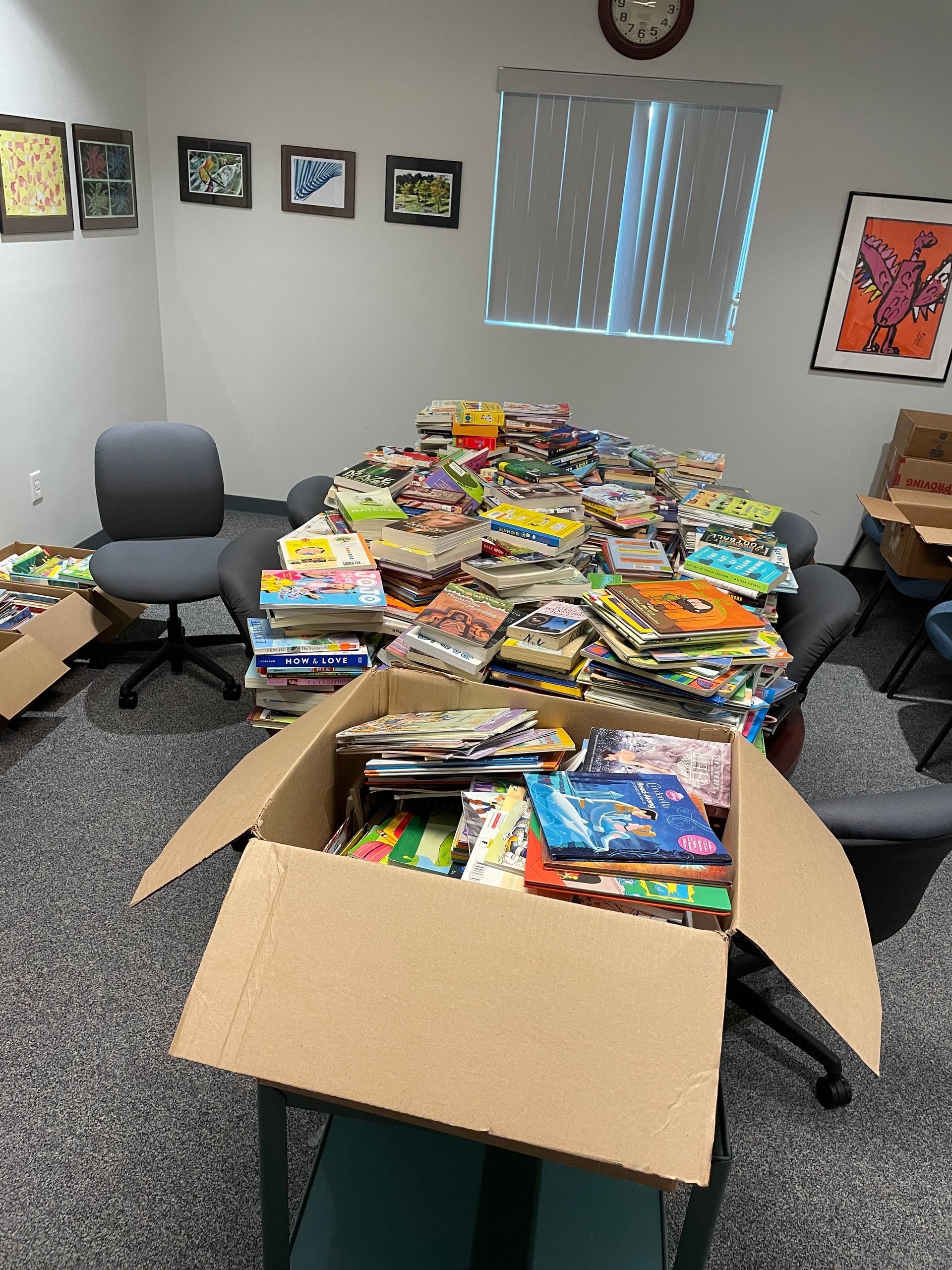 Books piled on a table and in a cardboard box, inside an office.