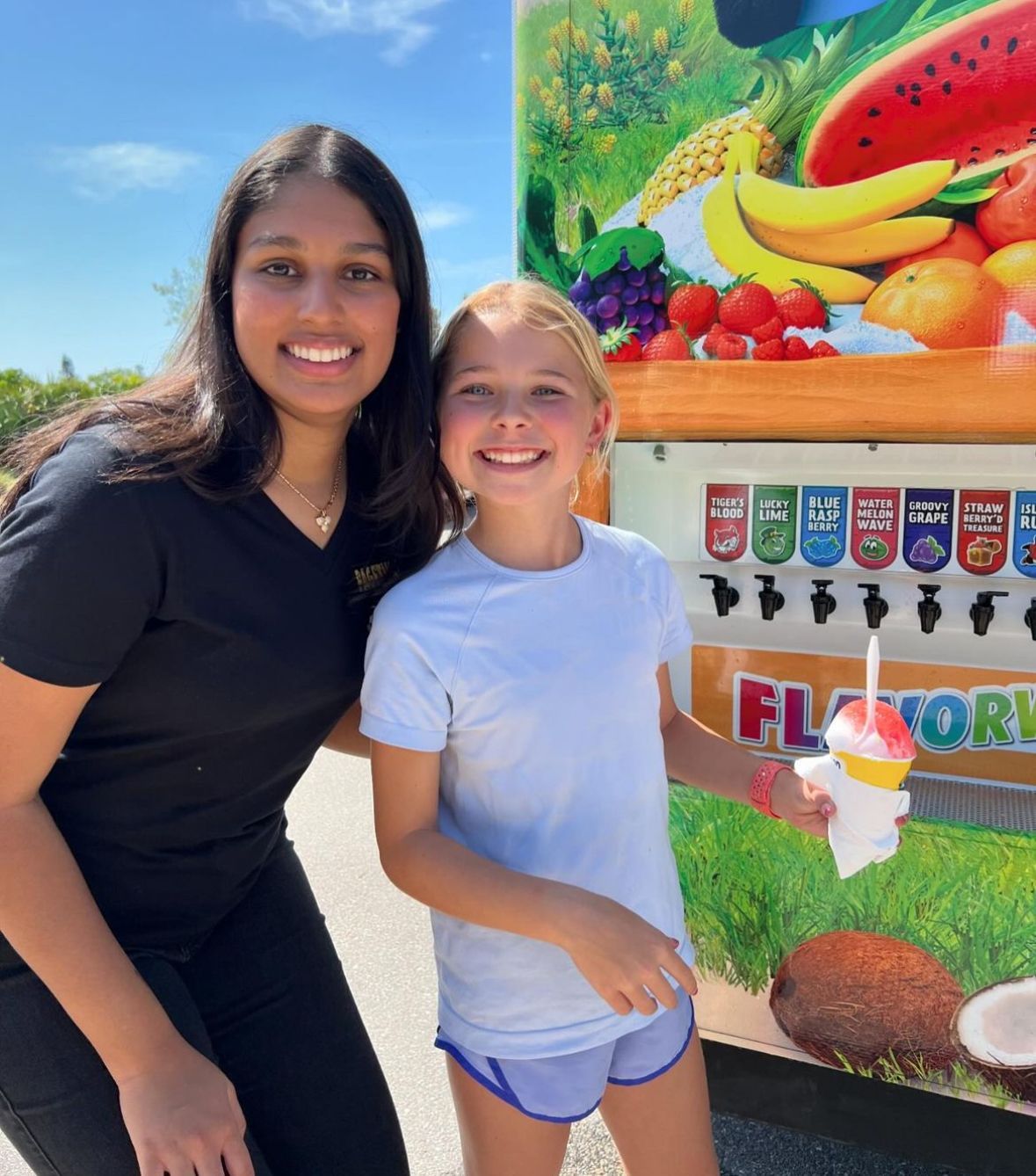 Woman and girl smile next to a flavor ice stand; girl holds ice. Sunny day.