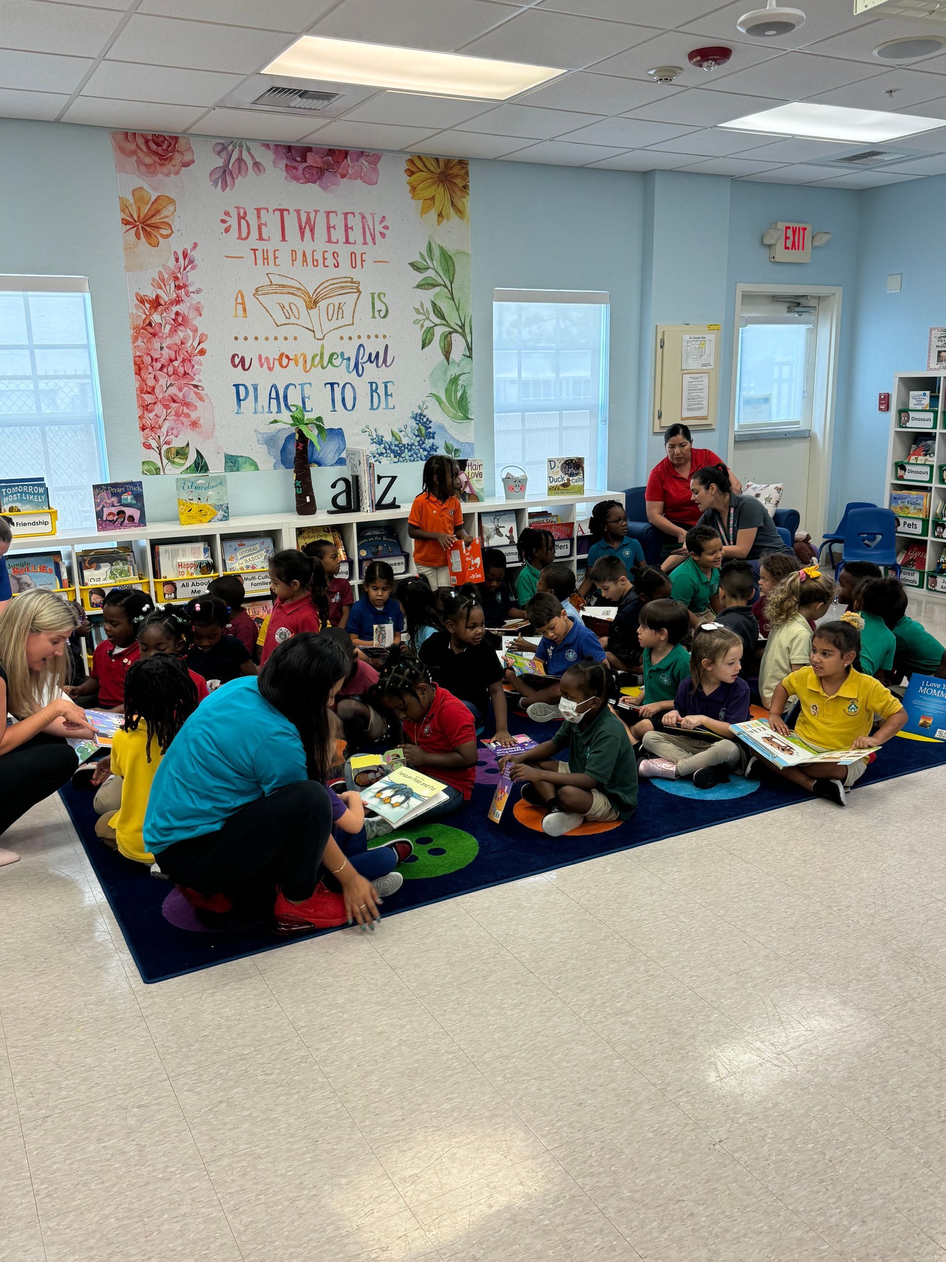 Children and teachers read in a classroom. Colorful mural on the wall; blue rug.