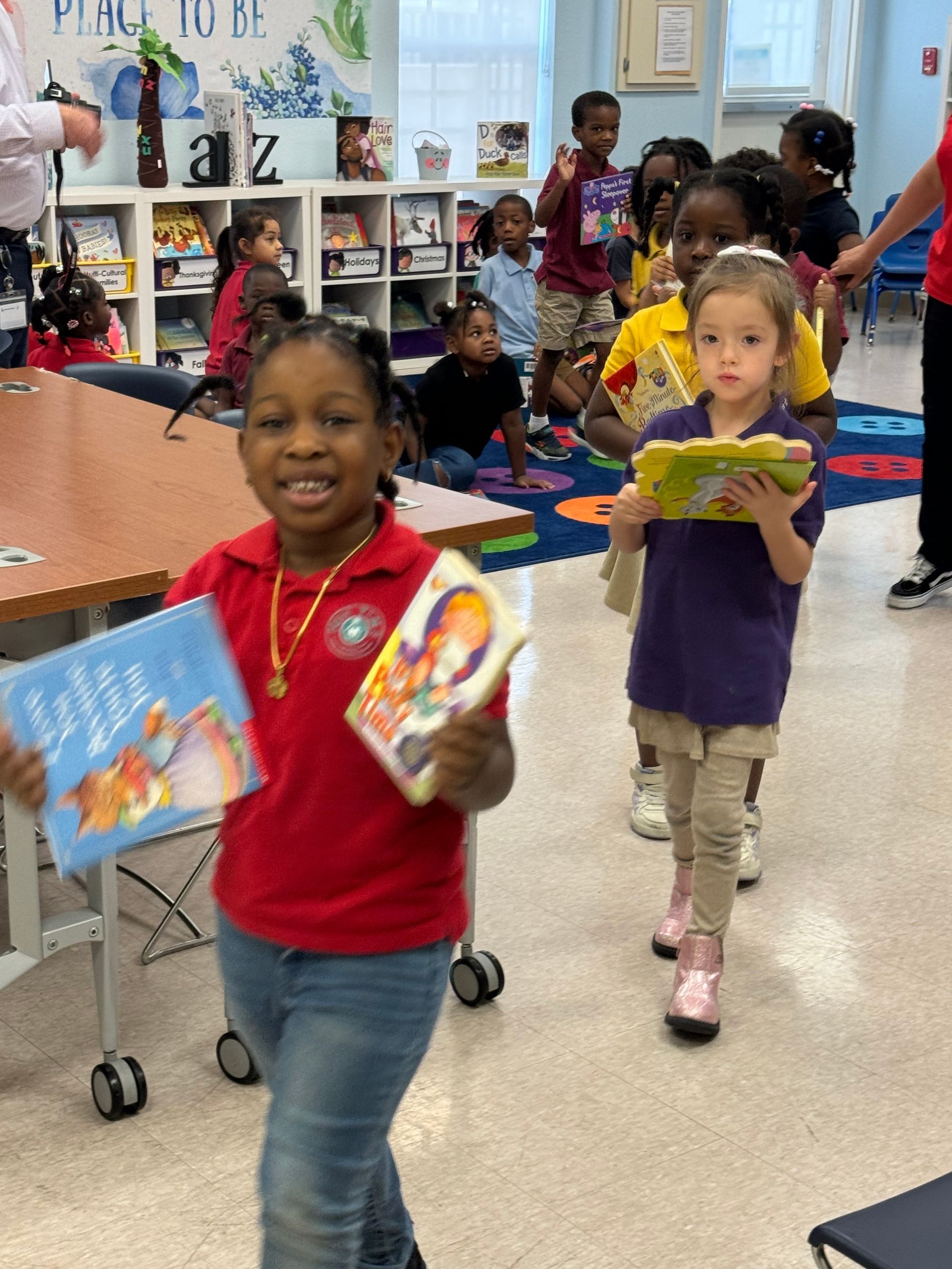 Children in classroom holding books, smiling. Some are walking, shelves with books in background.