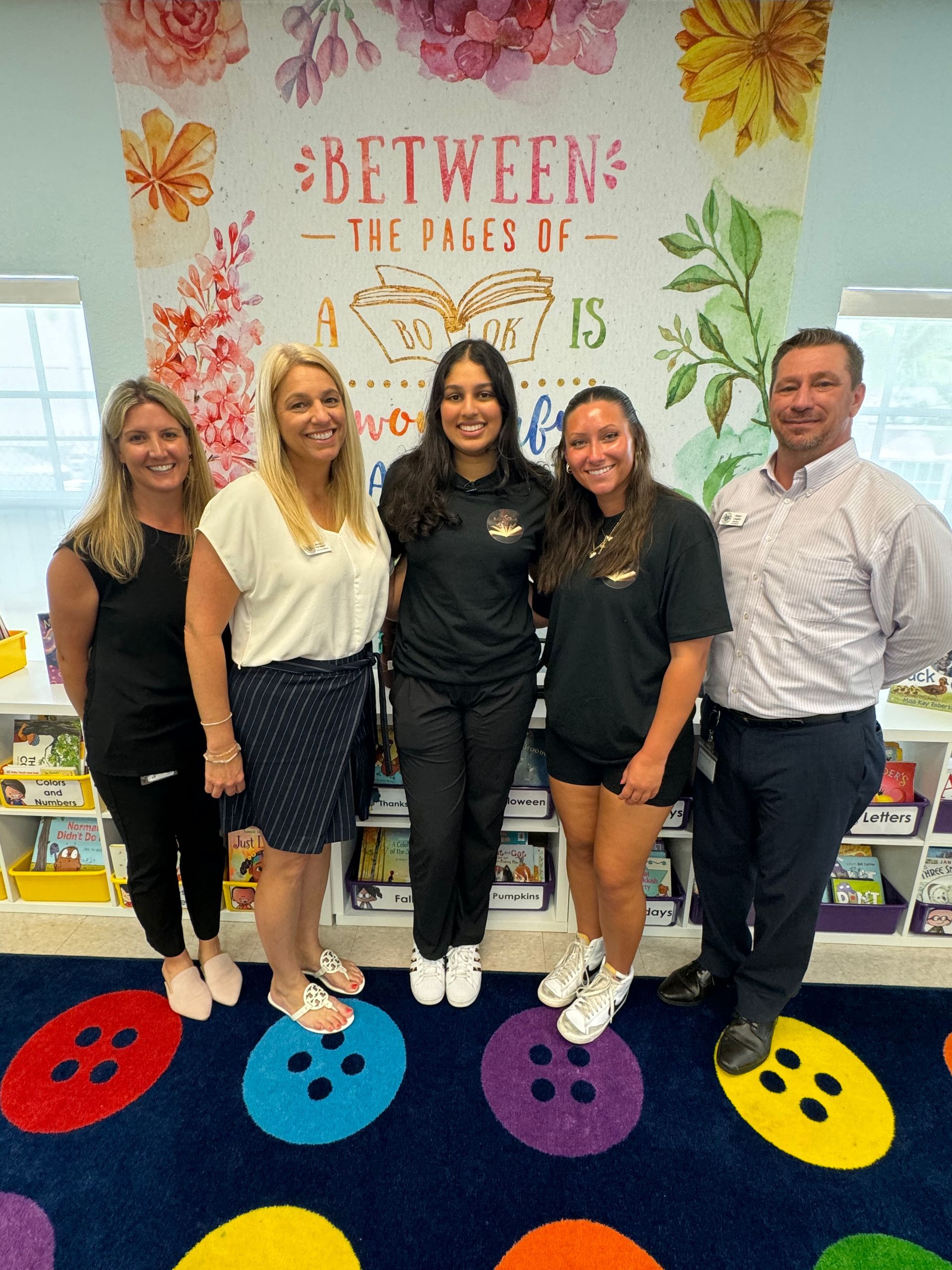 Five people stand in a colorful classroom. The background features a floral mural.