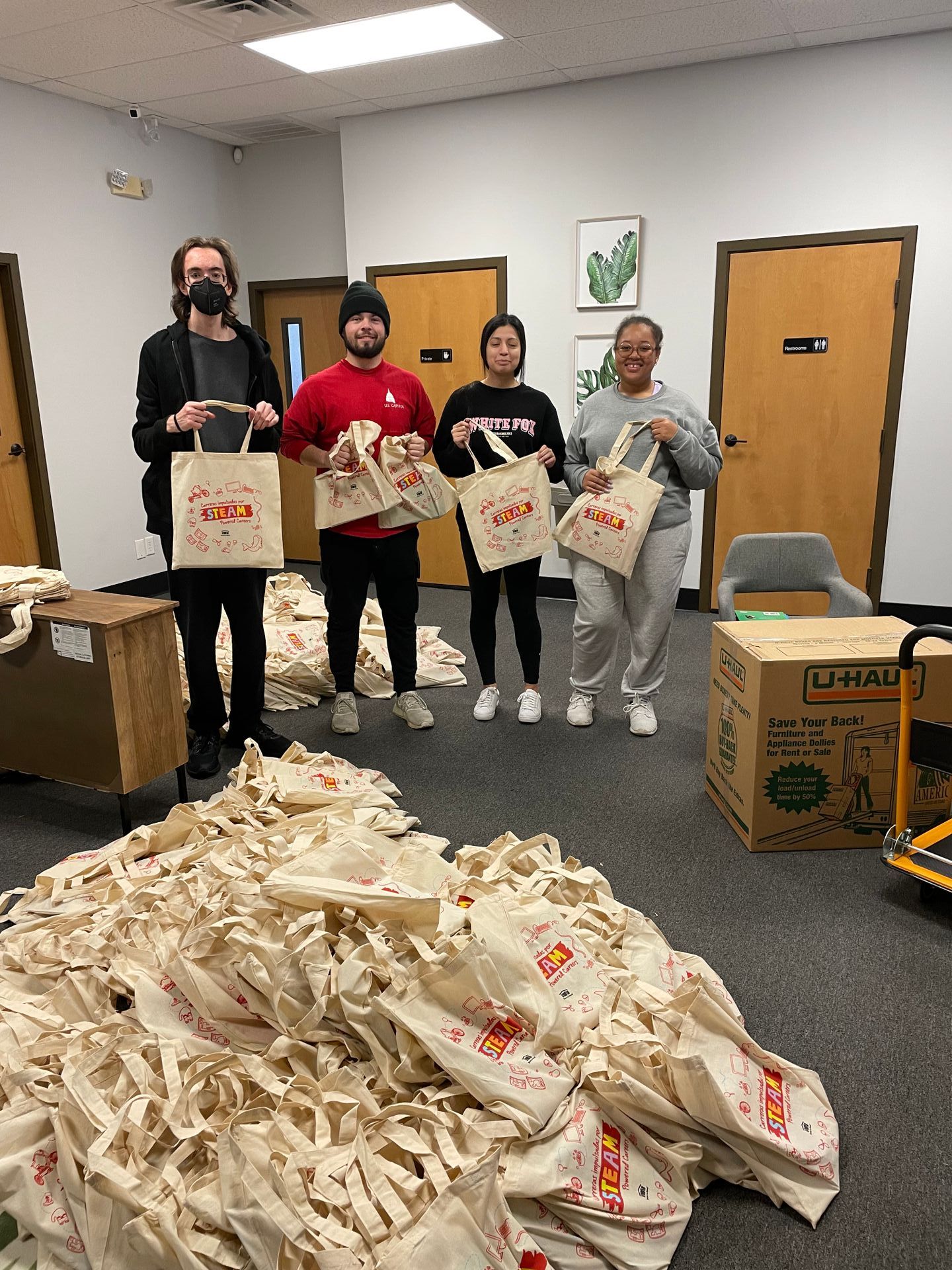 Four people stand with bags in a room, surrounded by piles of similar bags.