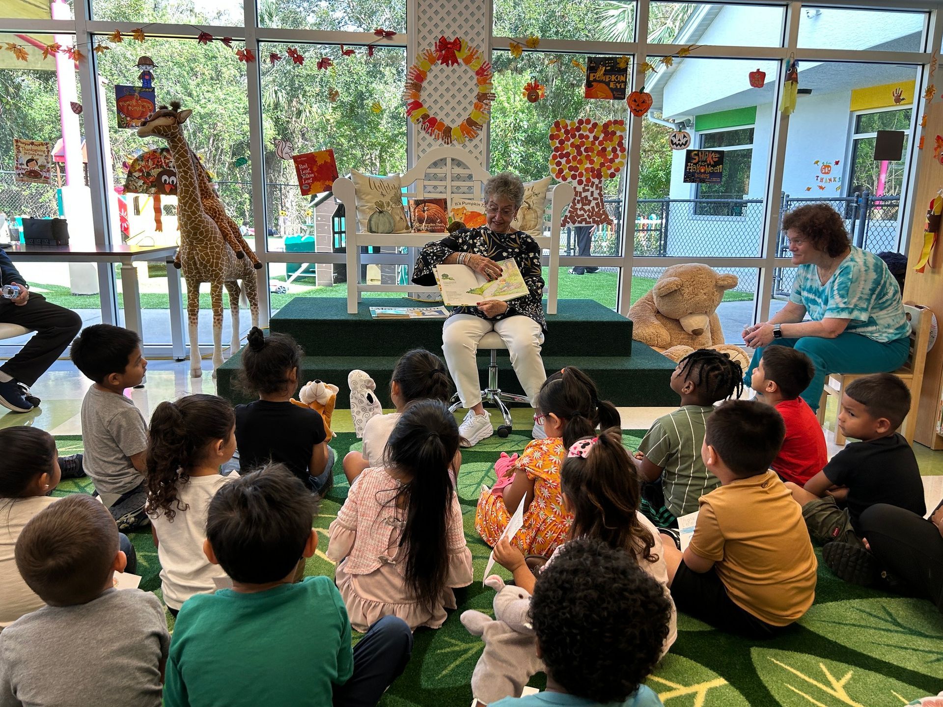 Children listen to a woman reading a book in a classroom, with fall decorations.