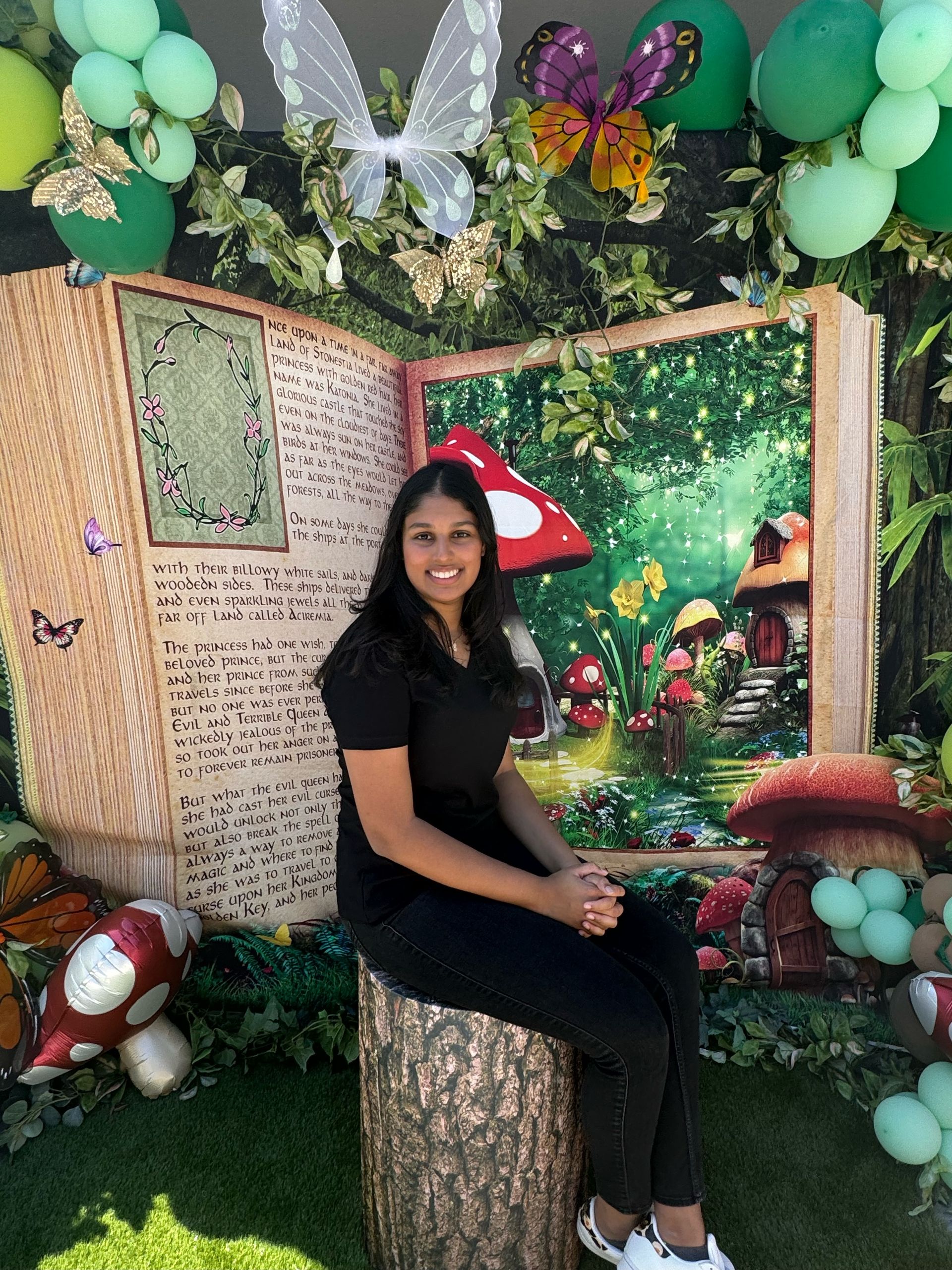 Woman sitting on a stump in front of a fairytale backdrop with balloons and decorations.