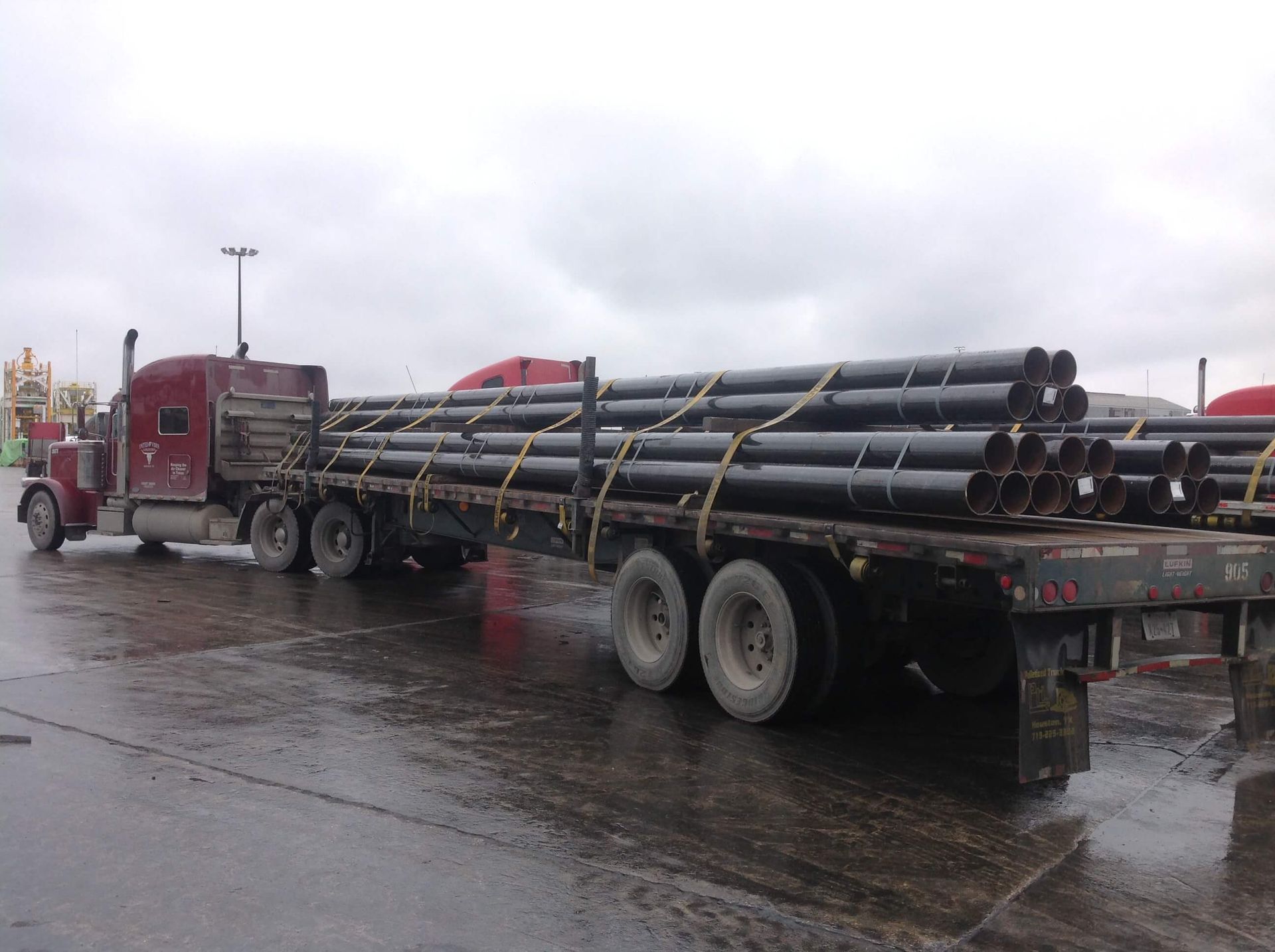 Semi-truck hauling a flatbed of long, black pipes on a wet, gray day.